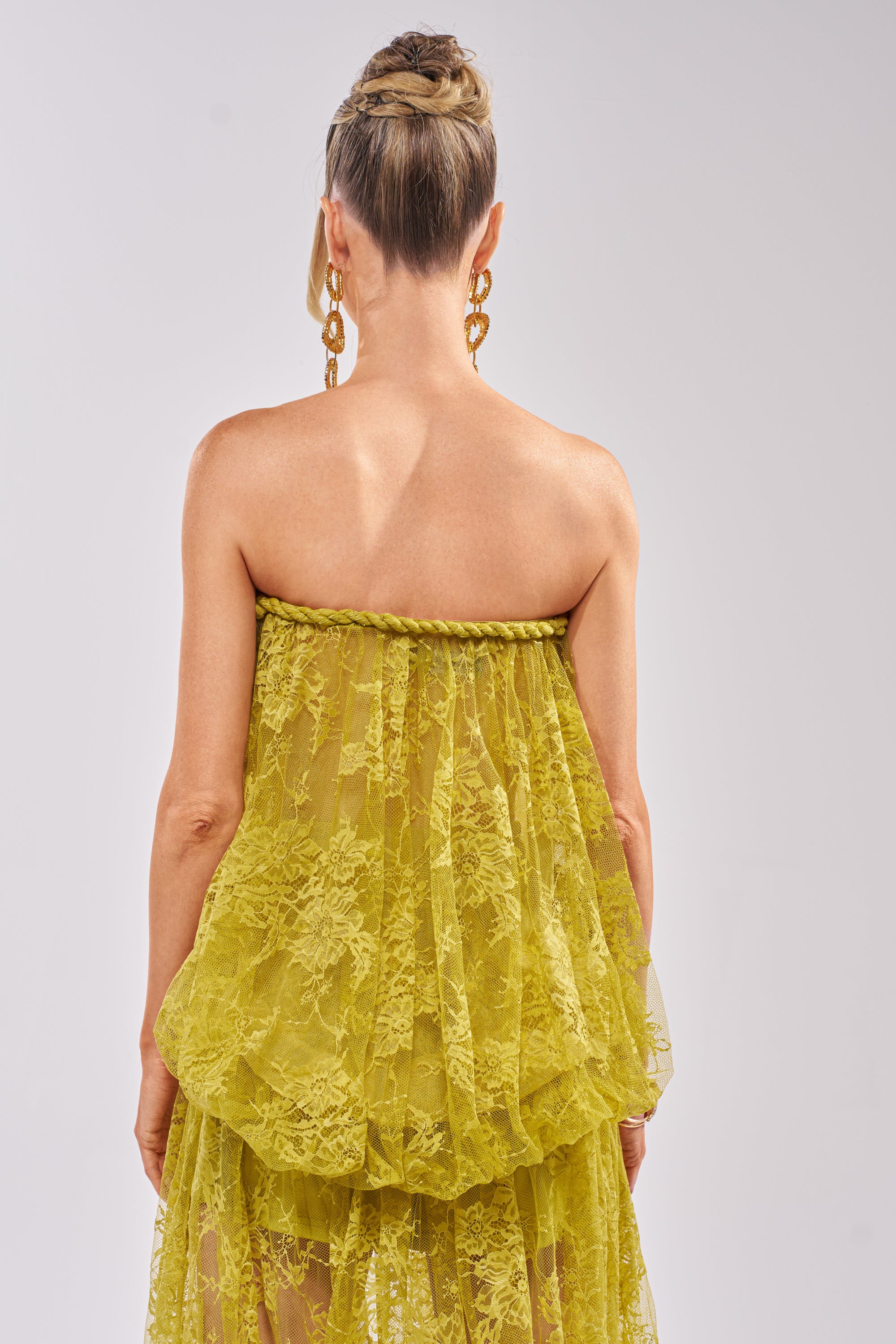 A woman with blonde hair in an updo is seen from behind, wearing the HANNAH LACE BLOUSE—a yellow strapless lace top with a sheer overlay—and large gold earrings, set against a plain light background. 5