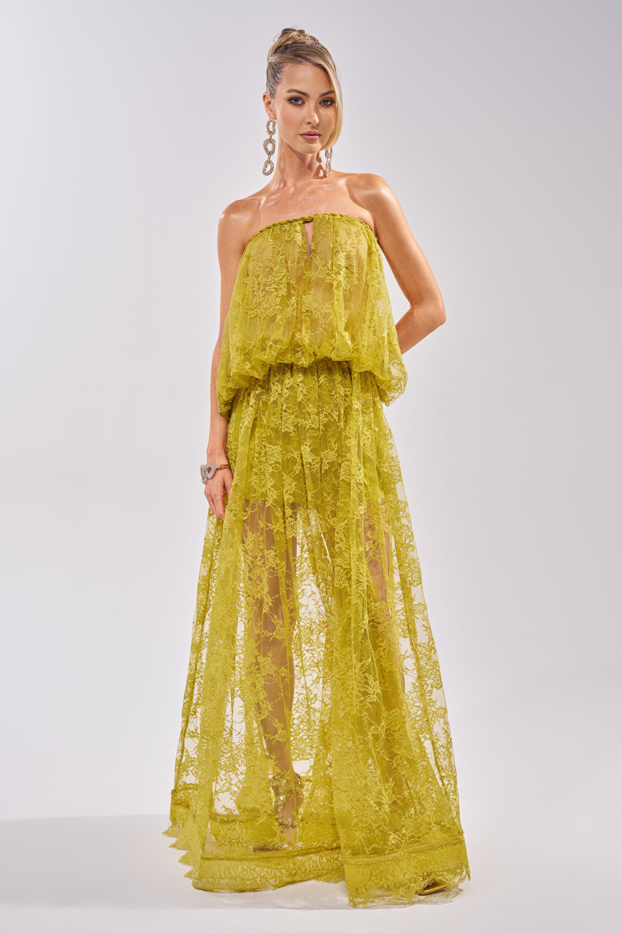 A woman poses against a plain background wearing the HANNAH LACE BLOUSE—a sheer, strapless yellow top with a loose, flowing silhouette. She accessorizes with long earrings and has her hair styled in an elegant updo. 7