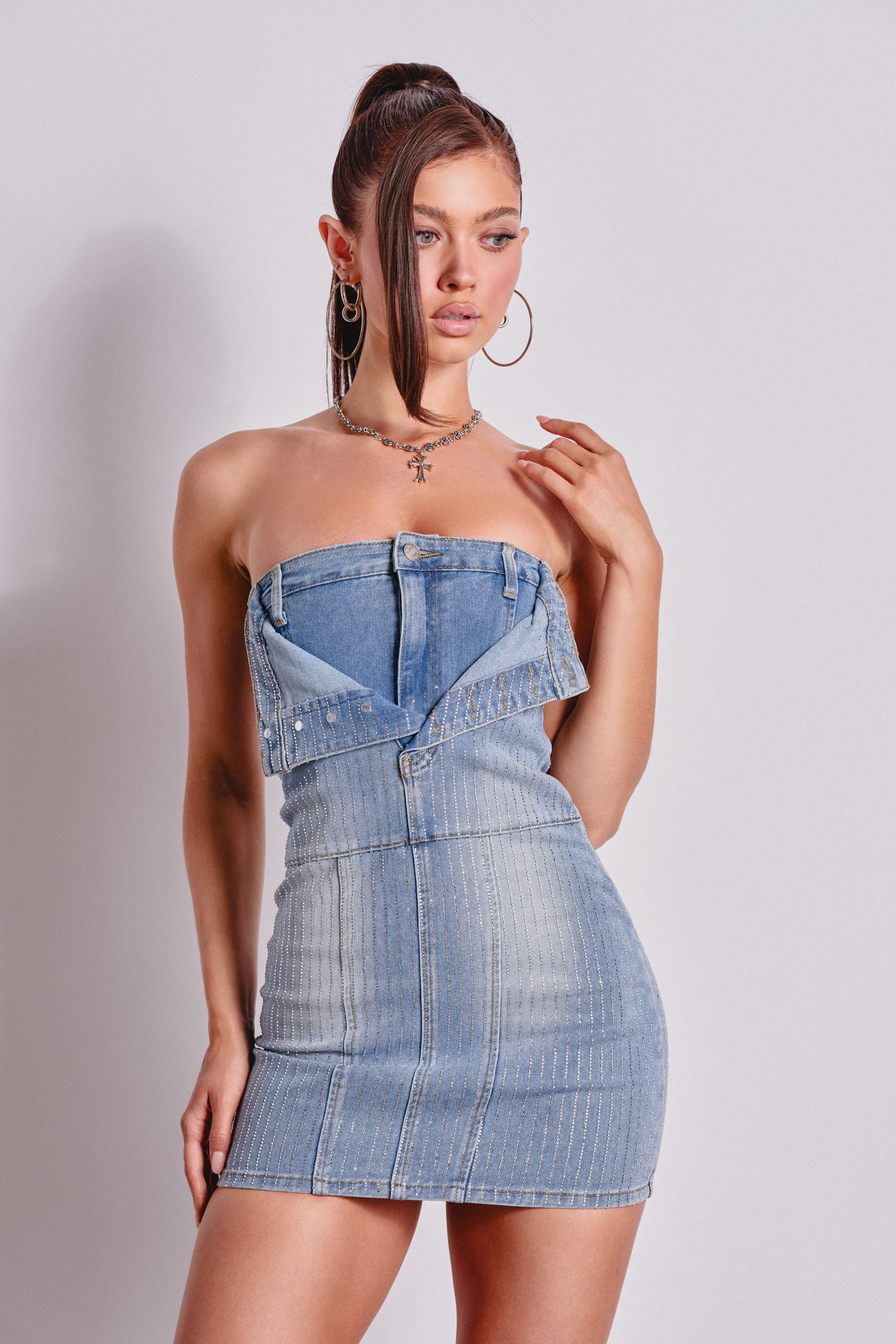 A woman poses against a plain background in the BUTTONED UP BEDAZZLED MINI DENIM DRESS, styled like an upside-down jean jacket. She finishes her streetwear look with hoop earrings, layered necklaces, and a high ponytail. 0