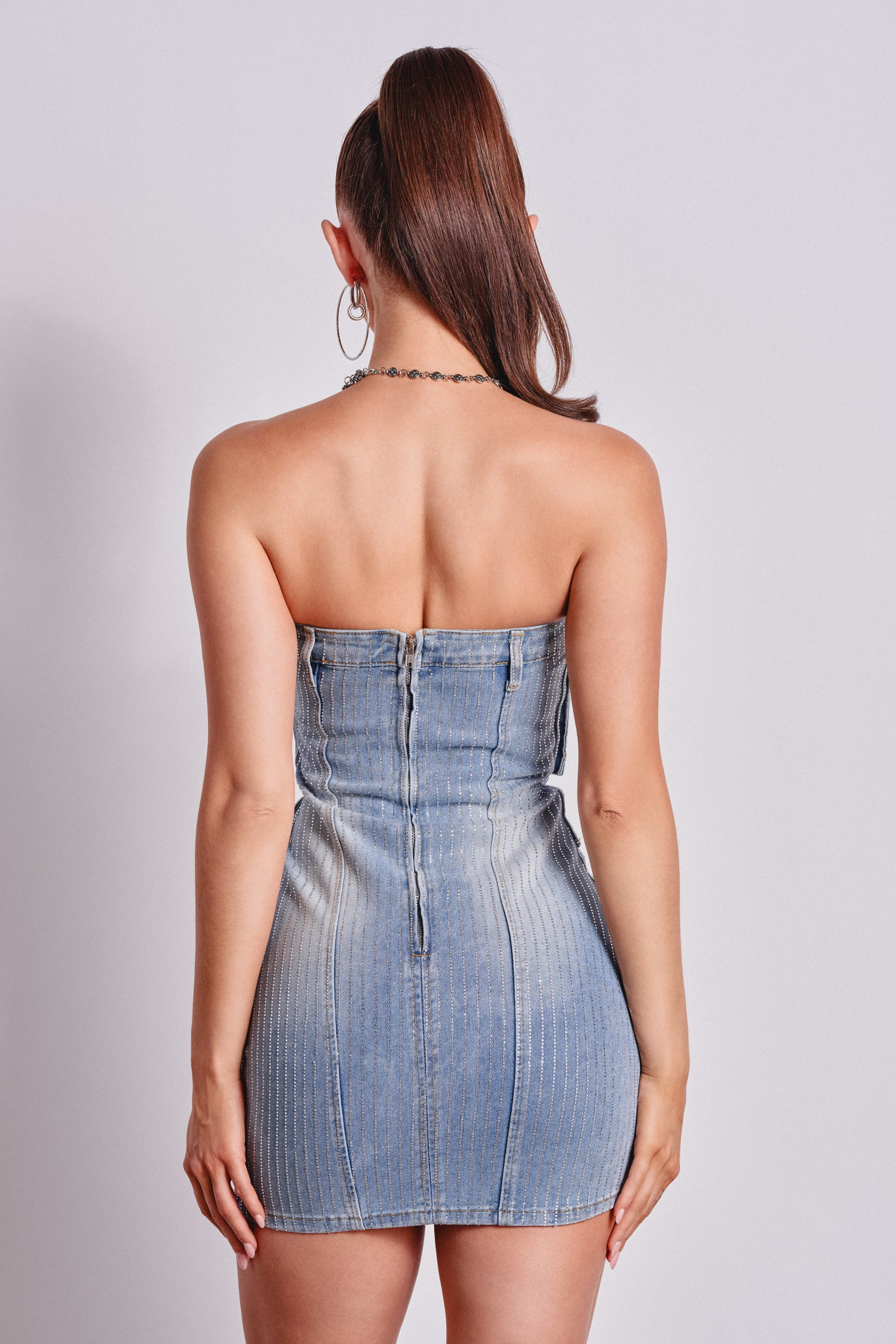 A woman with a high ponytail faces away, wearing the BUTTONED UP BEDAZZLED MINI DENIM DRESS with crystal embellishments. She stands against a plain light background, creating a chic look that's ideal for party outfits. 2