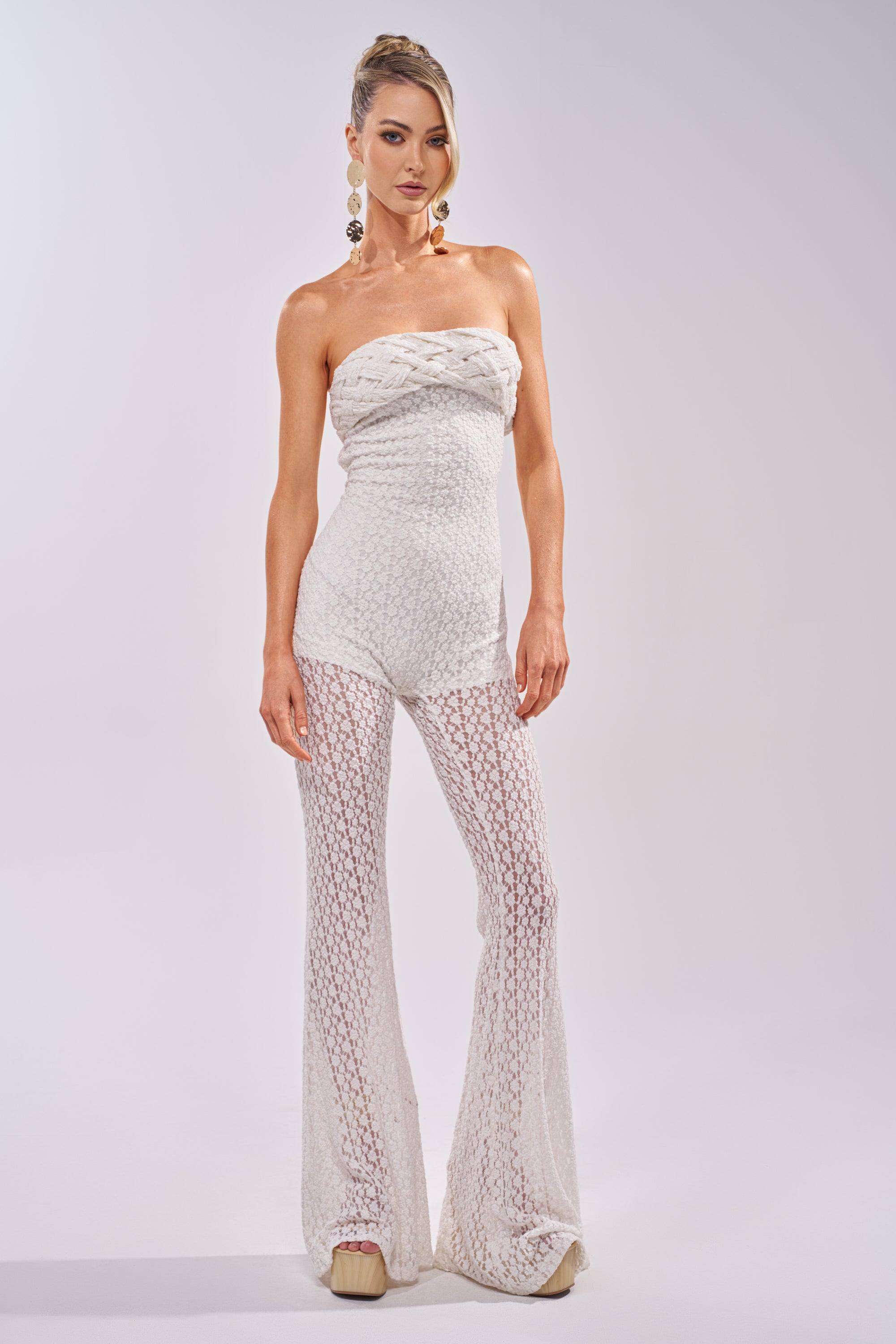 A woman stands against a light background wearing the CONCERT BRAT BRAIDED LACE JUMPSUIT IN WHITE, featuring strapless flared legs, nude platform shoes, and large gold earrings for a boho-chic look. 1