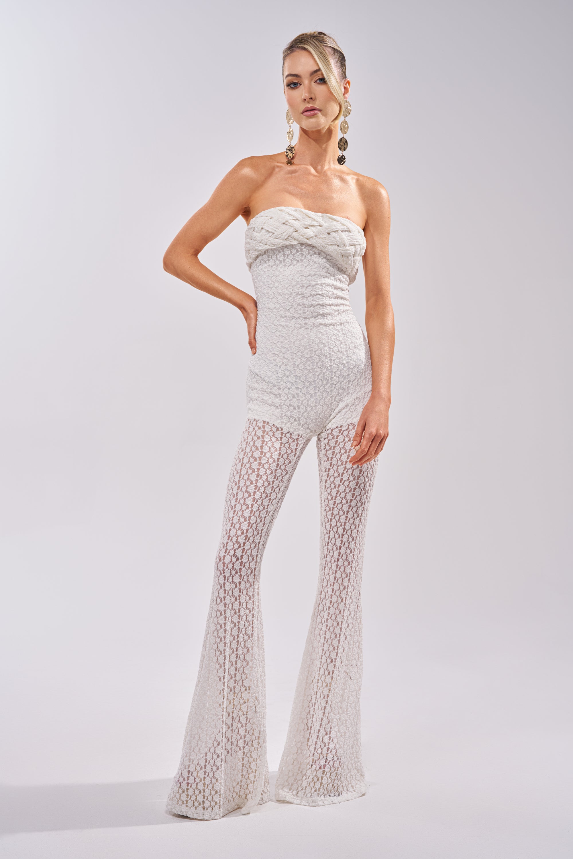 A woman poses confidently against a plain light background wearing the CONCERT BRAT BRAIDED LACE JUMPSUIT IN WHITE, featuring boho-chic style and flared legs. She accessorizes with long earrings and has her hair styled up. 3