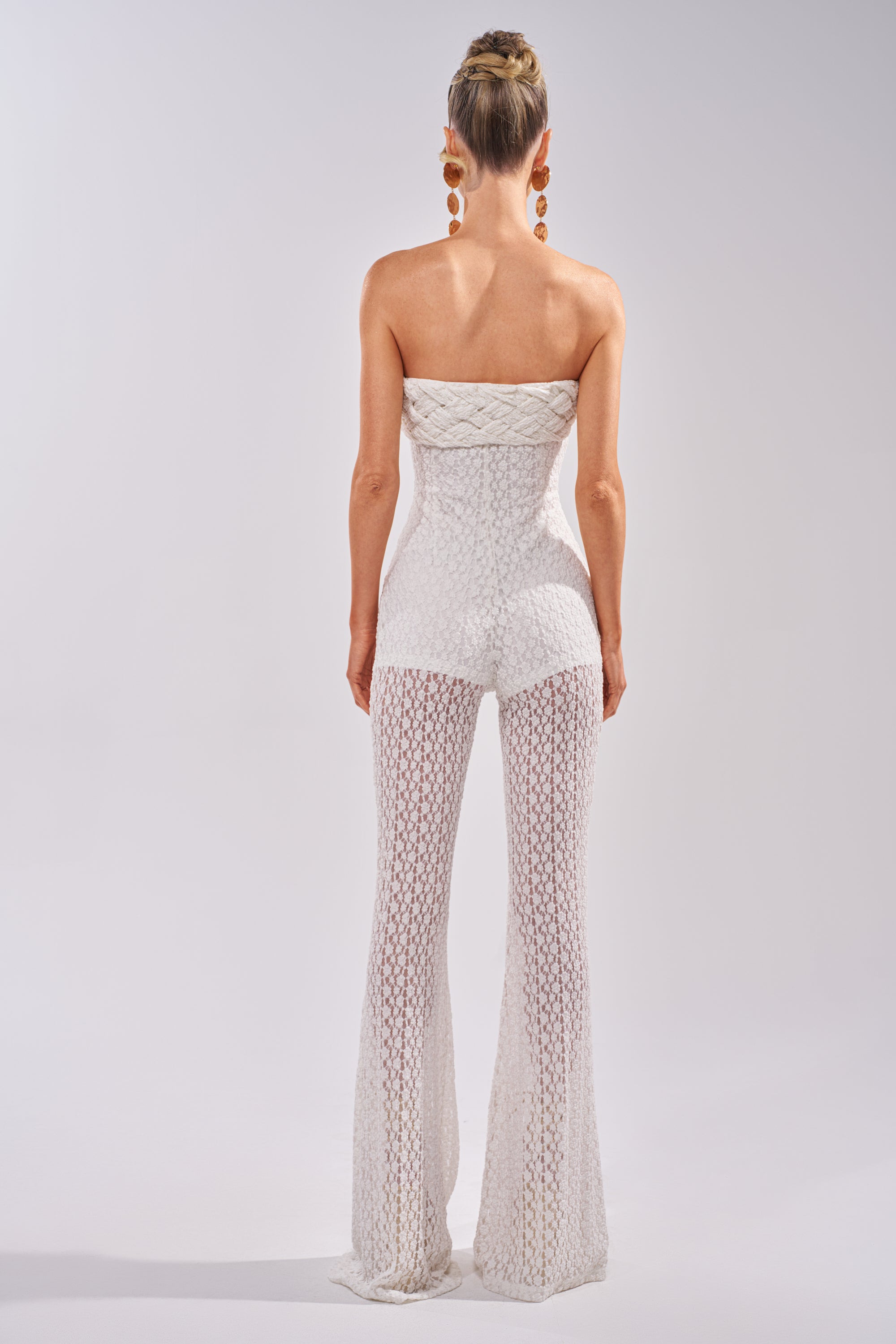 A woman with her hair in a bun models the CONCERT BRAT BRAIDED LACE JUMPSUIT IN WHITE, featuring flared legs, styled with large dangling earrings against a plain light background, facing away from the camera. 5