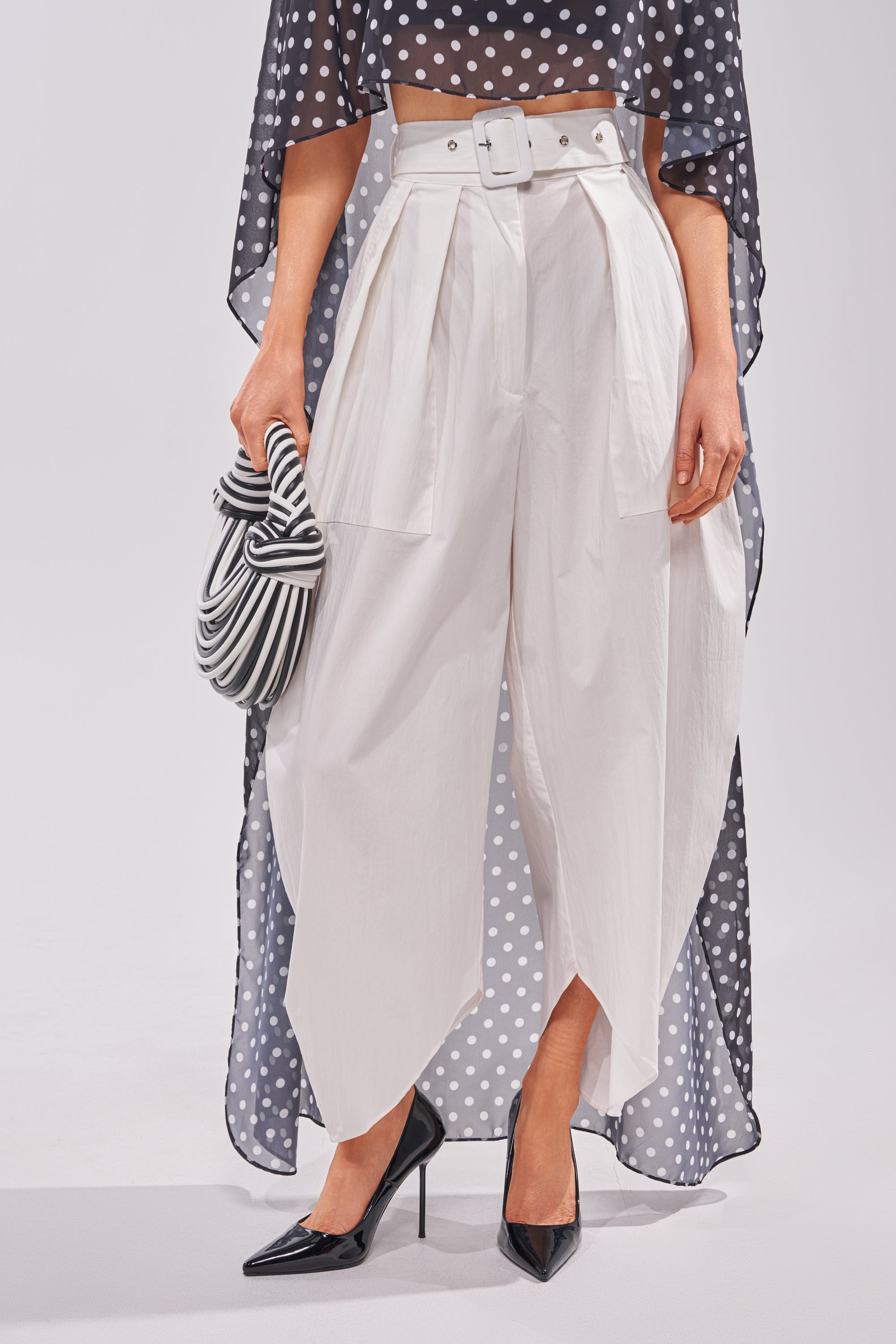 Someone showcases modern everyday style in the GIVE ME YOUR LOVE TROUSER—high-rise white barrel pants with an asymmetrical hem—paired with a white belt, polka dot top, black pointed heels, and a striped clutch. 0