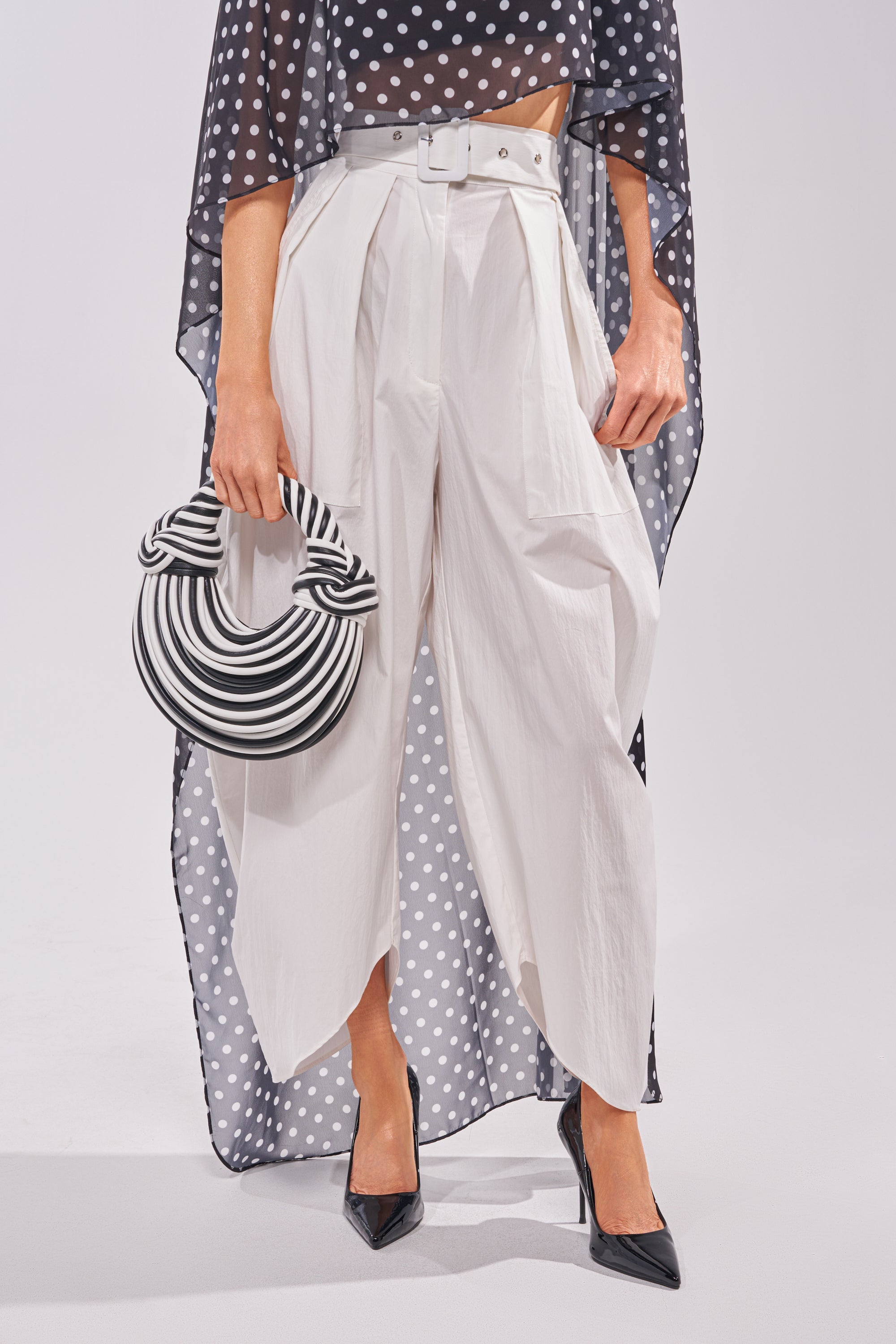 Showcasing modern style, a person wears the GIVE ME YOUR LOVE TROUSER in high-rise white, paired with a white belt, black pointed heels, a sheer black polka-dot top, and carries a round black-and-white striped handbag. Face not visible. 1