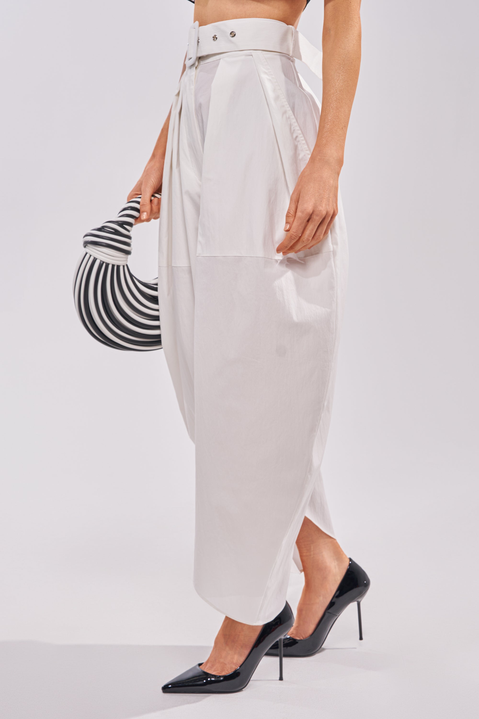 A woman wears the GIVE ME YOUR LOVE TROUSER, featuring high-rise, wide-leg white barrel pants with pleats, styled with black heels and a small black-and-white striped handbag. Only her lower half is visible. 2