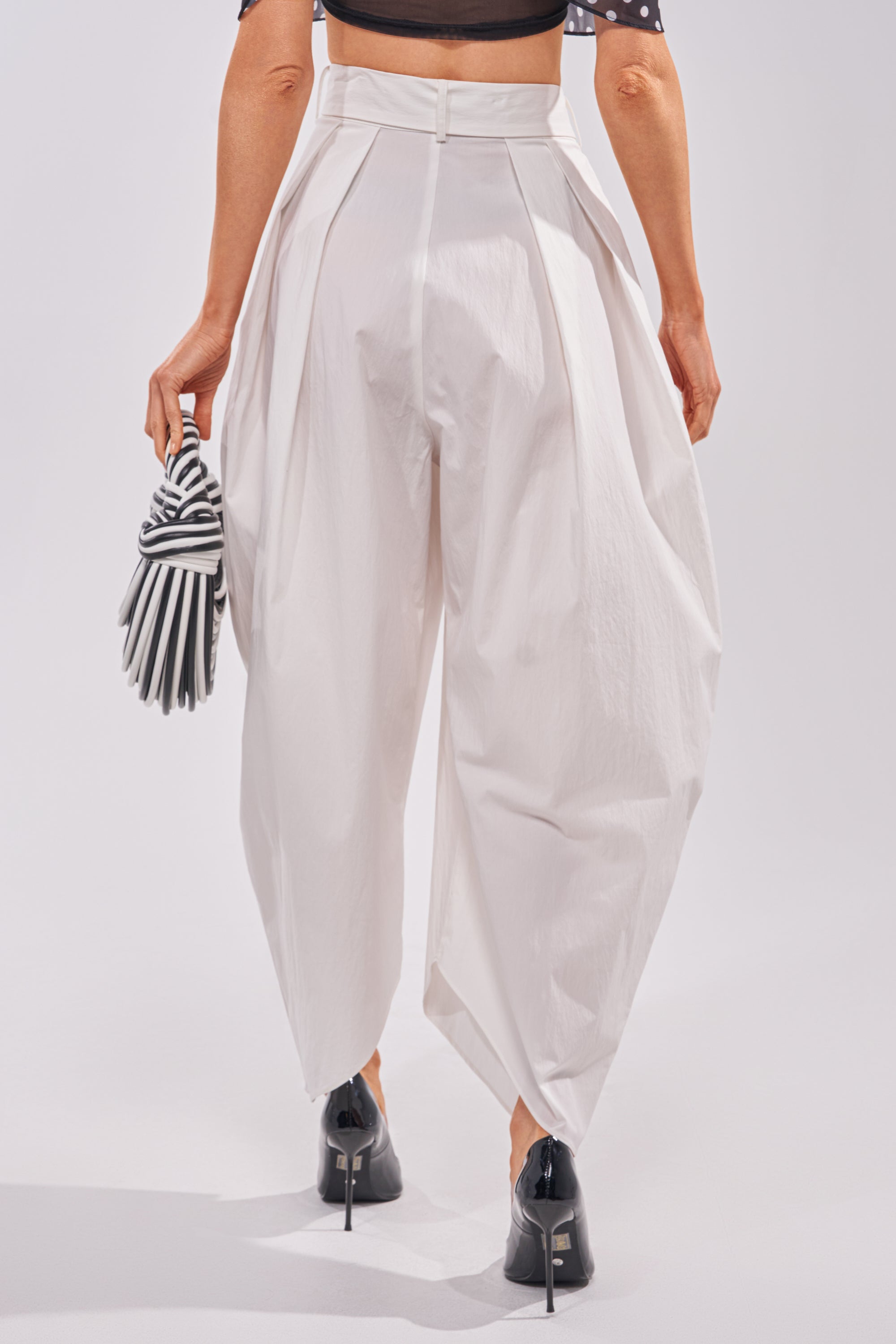 Wearing the GIVE ME YOUR LOVE TROUSER—high-rise, loose white barrel pants with pleats—a person pairs them with black heels, a black crop top, and a striped clutch. The photo is taken from behind, highlighting modern sculptural style. 3