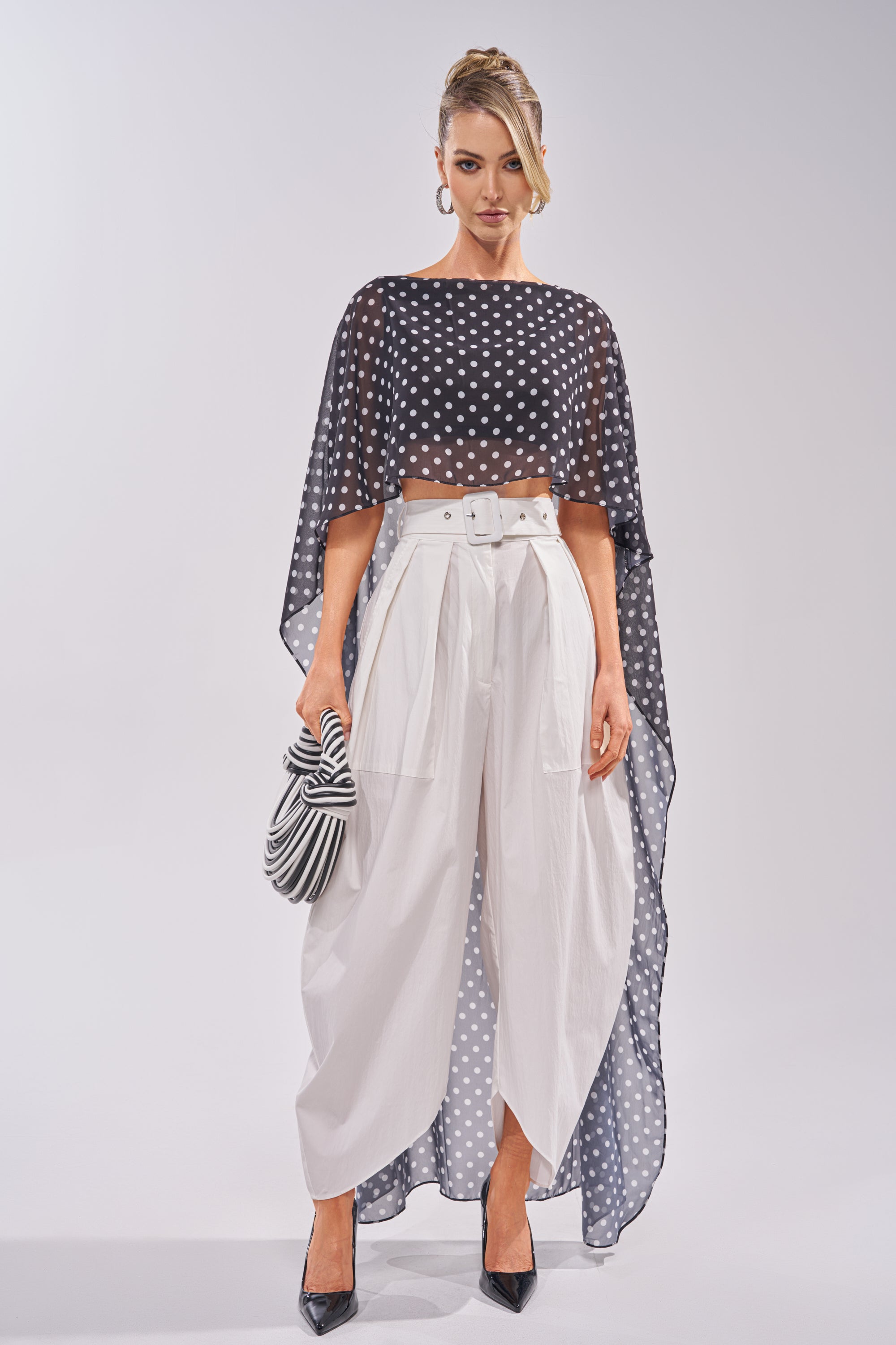 A woman stands wearing the GIVE ME YOUR LOVE TROUSER—high-rise, white barrel pants with a belt—styled with a sheer black polka-dot cape top, crop top, black heels, striped clutch, hoop earrings, and hair in a sculptural updo. 4