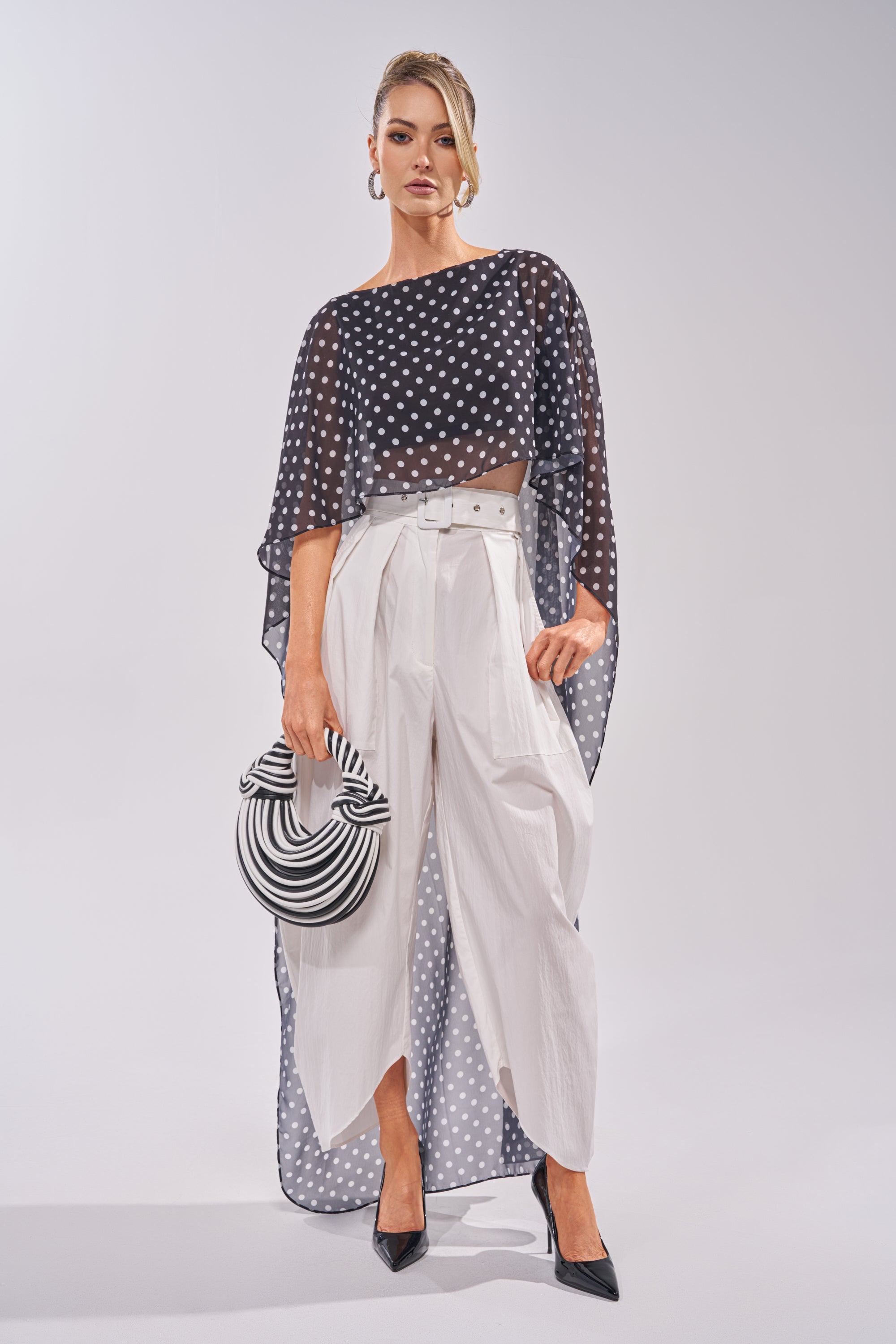A woman stands confidently in sheer black polka-dot top over a black crop top, GIVE ME YOUR LOVE TROUSER in white with a belt, black heels, hoop earrings, and holding a black-and-white striped hat. 5