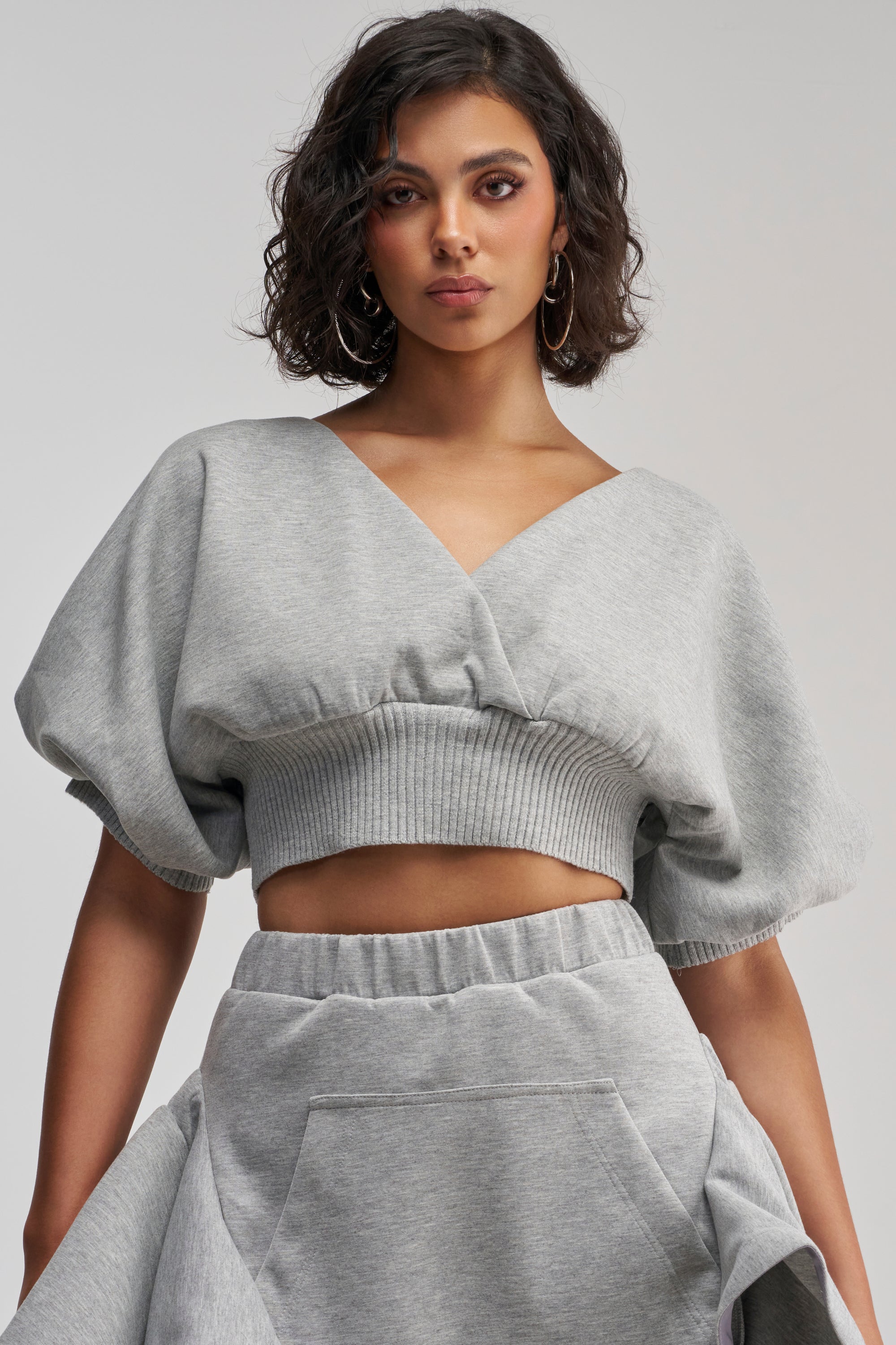 A woman with wavy dark hair wears the NO WORK ALL PLAY SCUBA SWEATSHIRT in light gray, styled with a matching high-waisted skirt. She stands against a neutral background, looking confidently at the camera.
