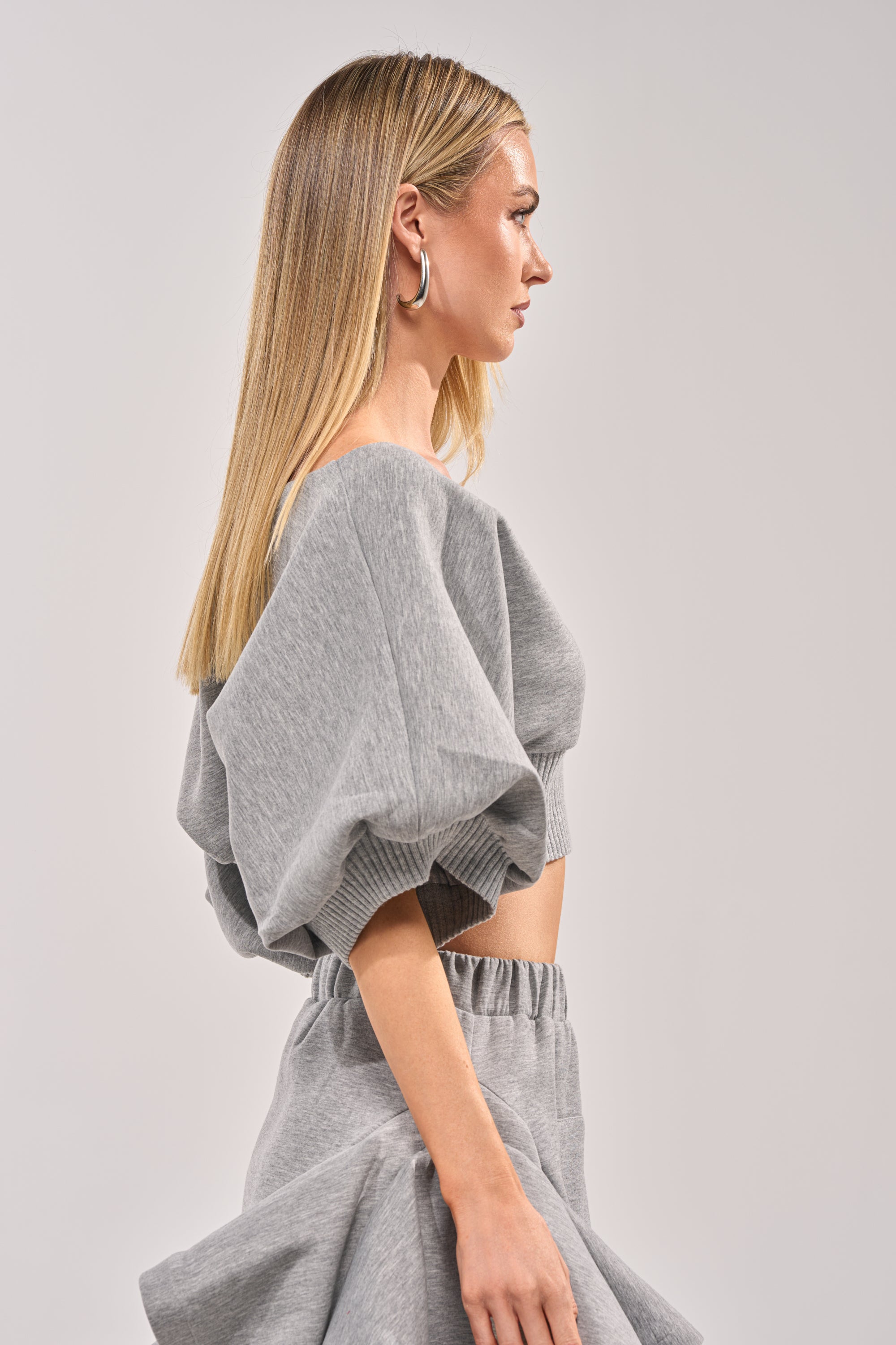 A woman with straight blonde hair models the NO WORK ALL PLAY SCUBA SWEATSHIRT, a grey, cropped streetwear top with puff sleeves, paired with a matching skirt. She is shown in profile against a plain light background. 3