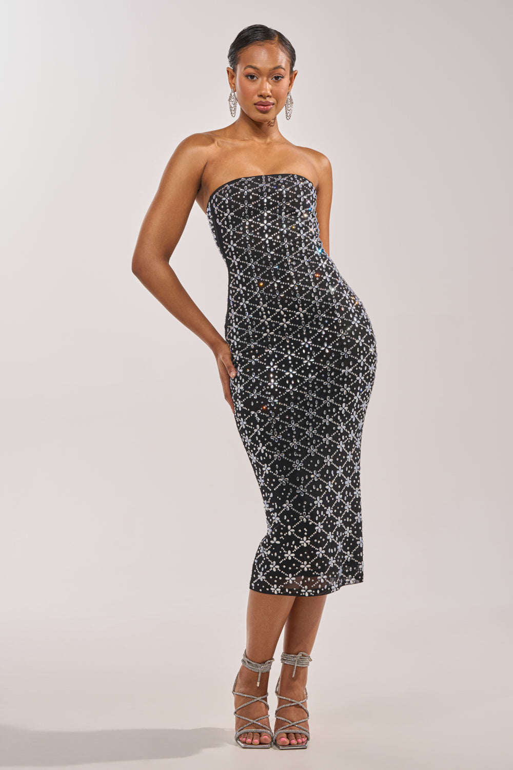 A woman poses against a plain light background in the MORE THE MERRIER EMBELLISHED MAXI DRESS—strapless, fitted, with a white geometric pattern—paired with silver strappy heels and large hoop earrings. 1
