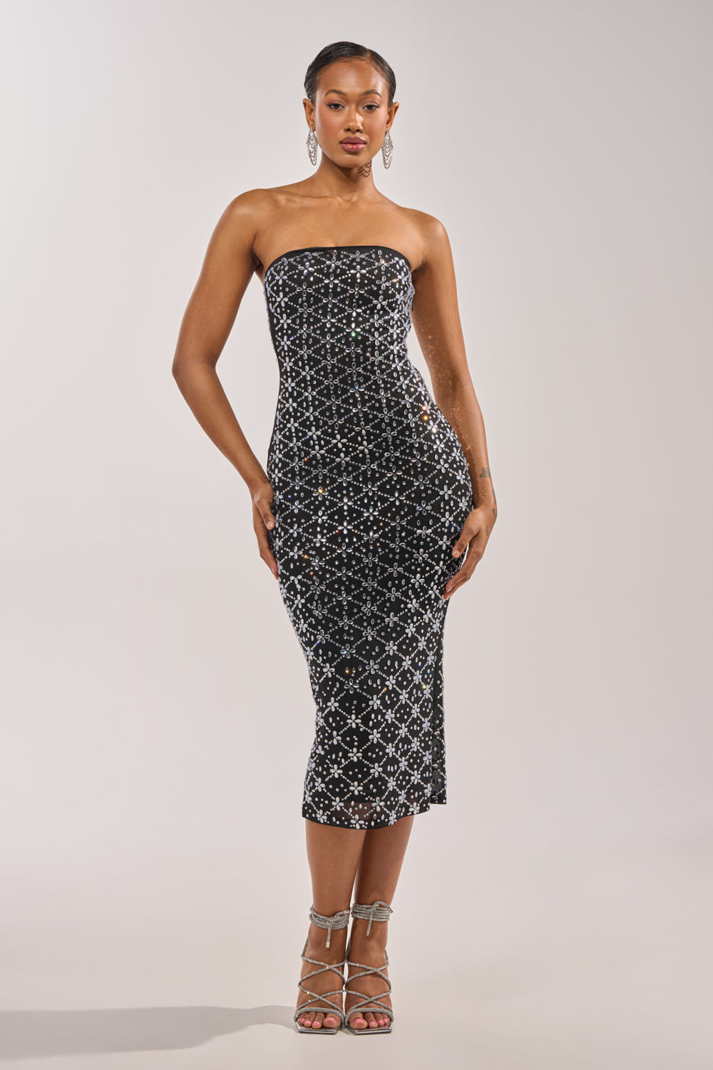 A woman models the MORE THE MERRIER EMBELLISHED MAXI DRESS—a black, form-fitting strapless dress with silver geometric sequins—paired with strappy high heels. Her hair is styled back as she stands against a simple light background. 3