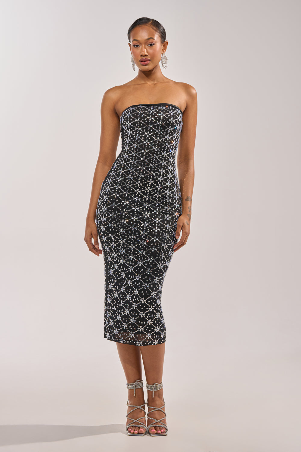 A woman stands against a plain light background in the MORE THE MERRIER EMBELLISHED MAXI DRESS, a strapless, body-contouring black dress with white and silver sequin details, paired with silver strappy heels. 4