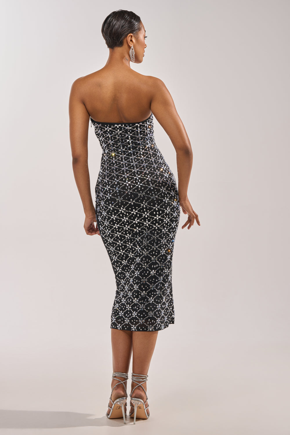 A woman faces away in the MORE THE MERRIER EMBELLISHED MAXI DRESS, a body-contouring formal piece with sparkling silver geometric patterns and silver strappy heels. Her sleek short hair completes the striking look against a plain light background. 6