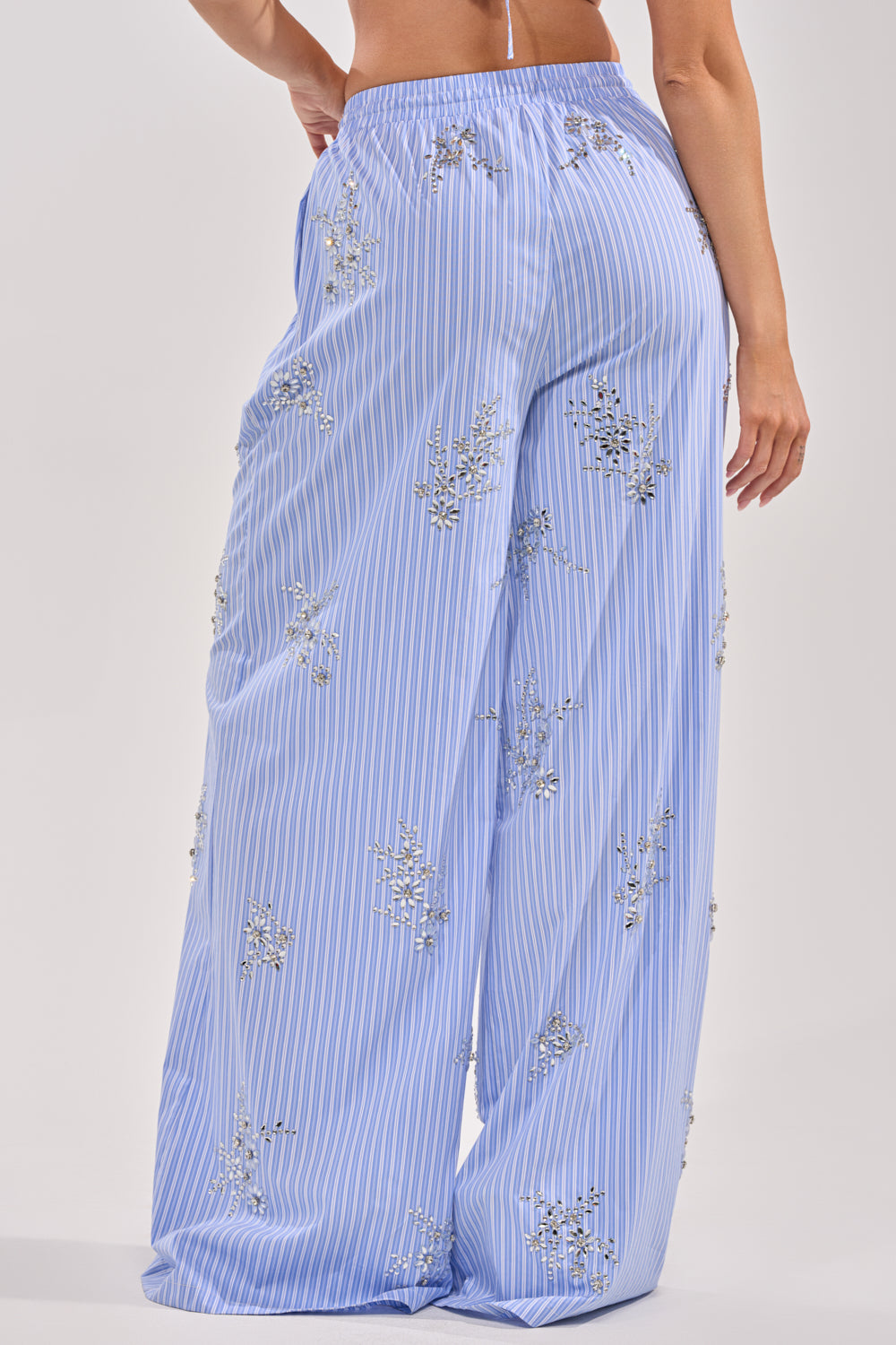 A person is seen from the back wearing SWEET DREAMS RHINESTONE POPLIN PANT, featuring white vertical stripes, delicate floral embroidery, and a resort-inspired flowy fit. 5