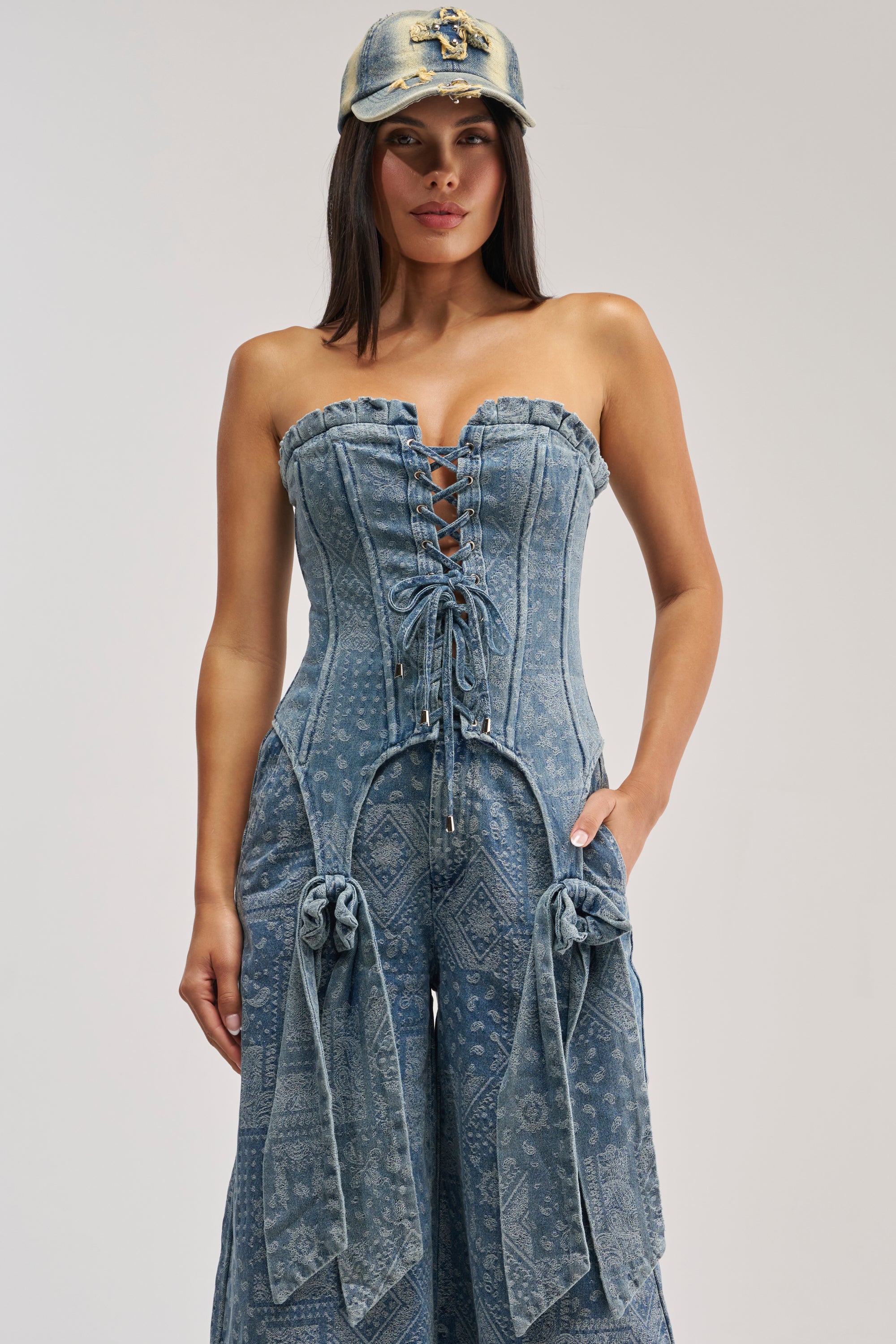 A woman wearing the DREAMING BIG PAISLEY DENIM CORSET—a strapless, lace-up corset with bow details and wide legs—paired with a distressed denim baseball cap, stands against a plain background.