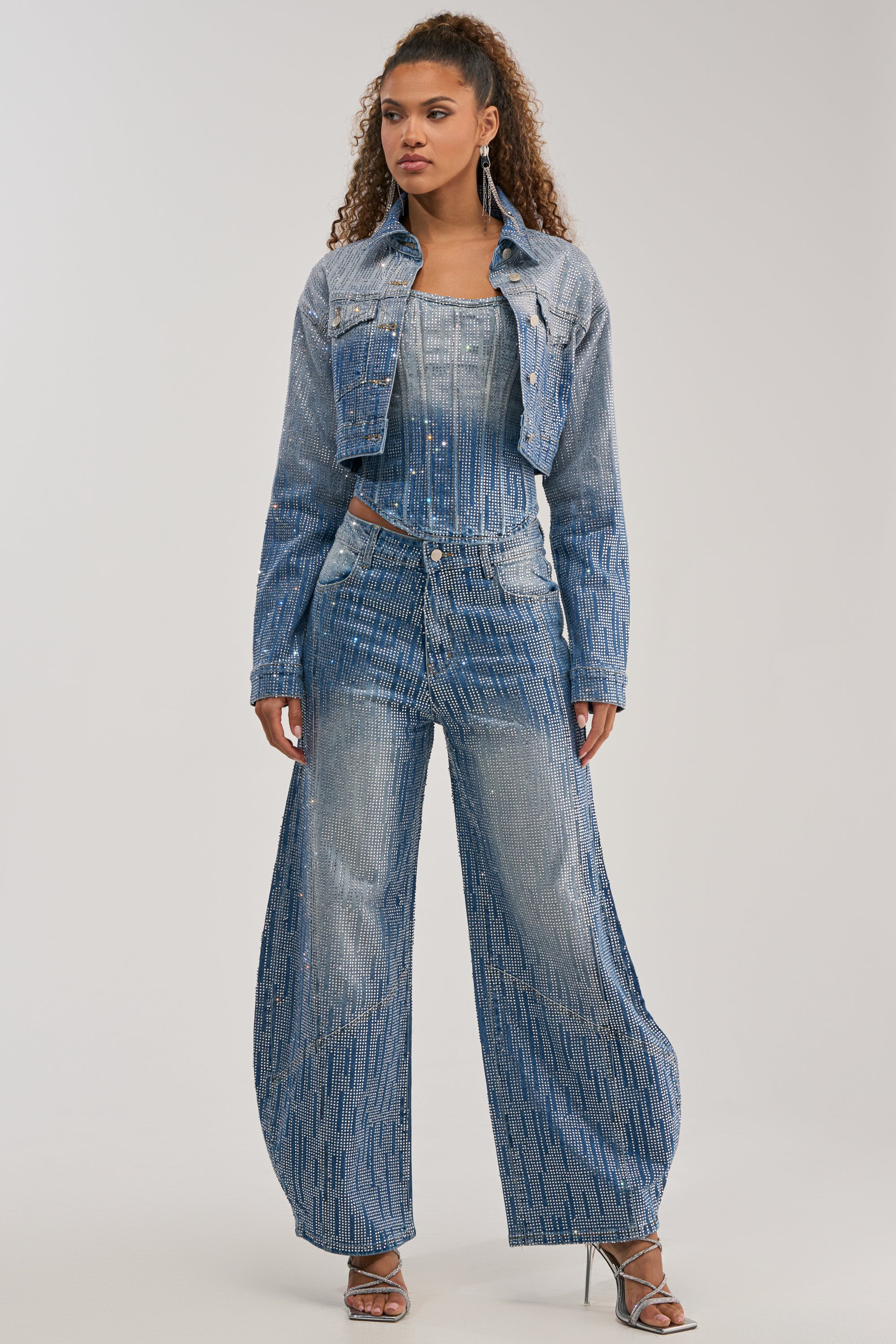 A woman stands against a plain light background wearing the MIND SPINNING EMBELLISHED DENIM set—a coordinated denim jacket, crop top, and wide-leg jeans featuring rhinestone ombre denim embellishments—paired with strappy silver heels for a striking statement streetwear look.