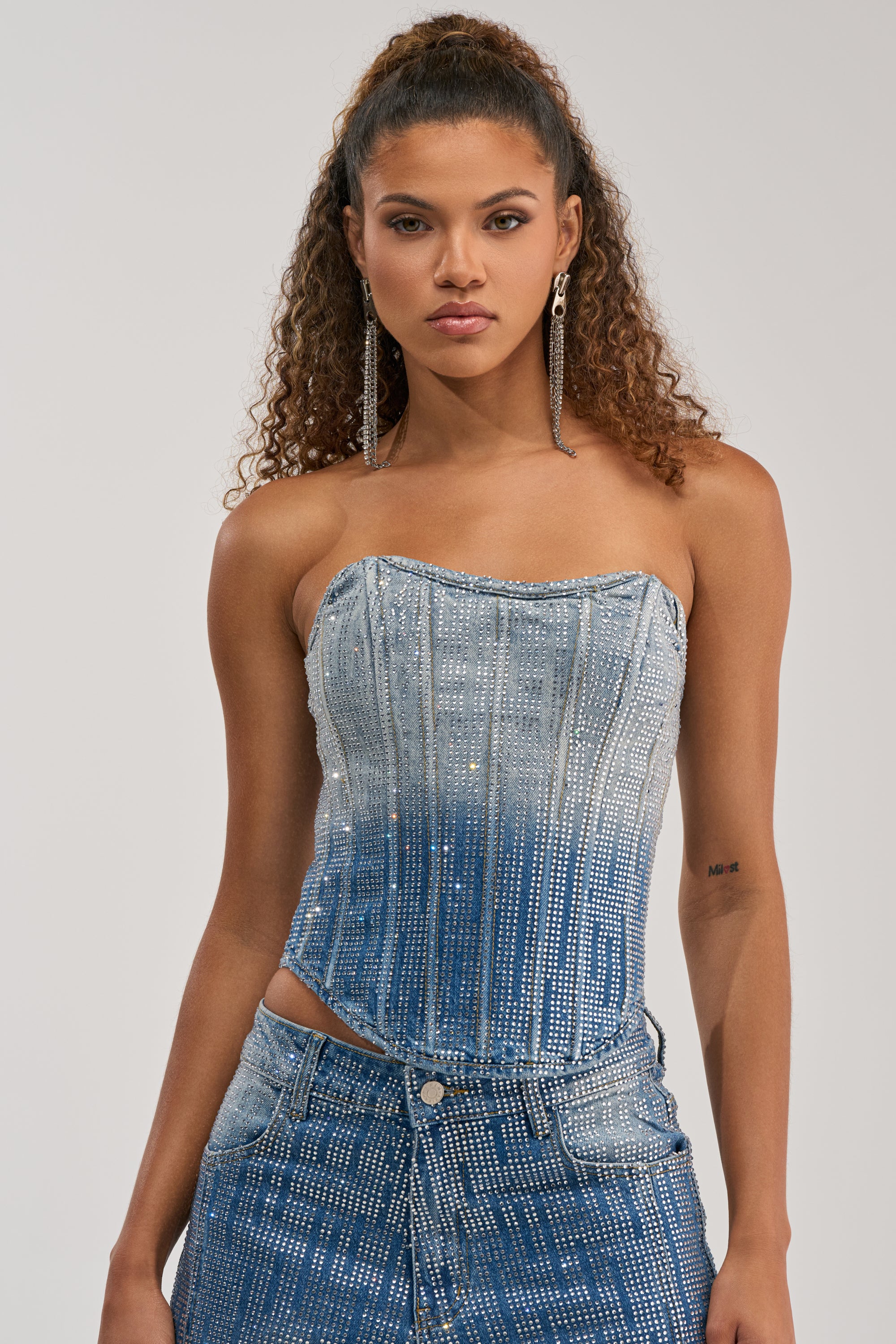 A woman with curly hair wears the MIND SPINNING EMBELLISHED DENIM CORSET and matching denim pants, both adorned with sparkly rhinestones. She pairs her look with long earrings and poses confidently against a plain light background—an effortlessly chic take on statement streetwear.