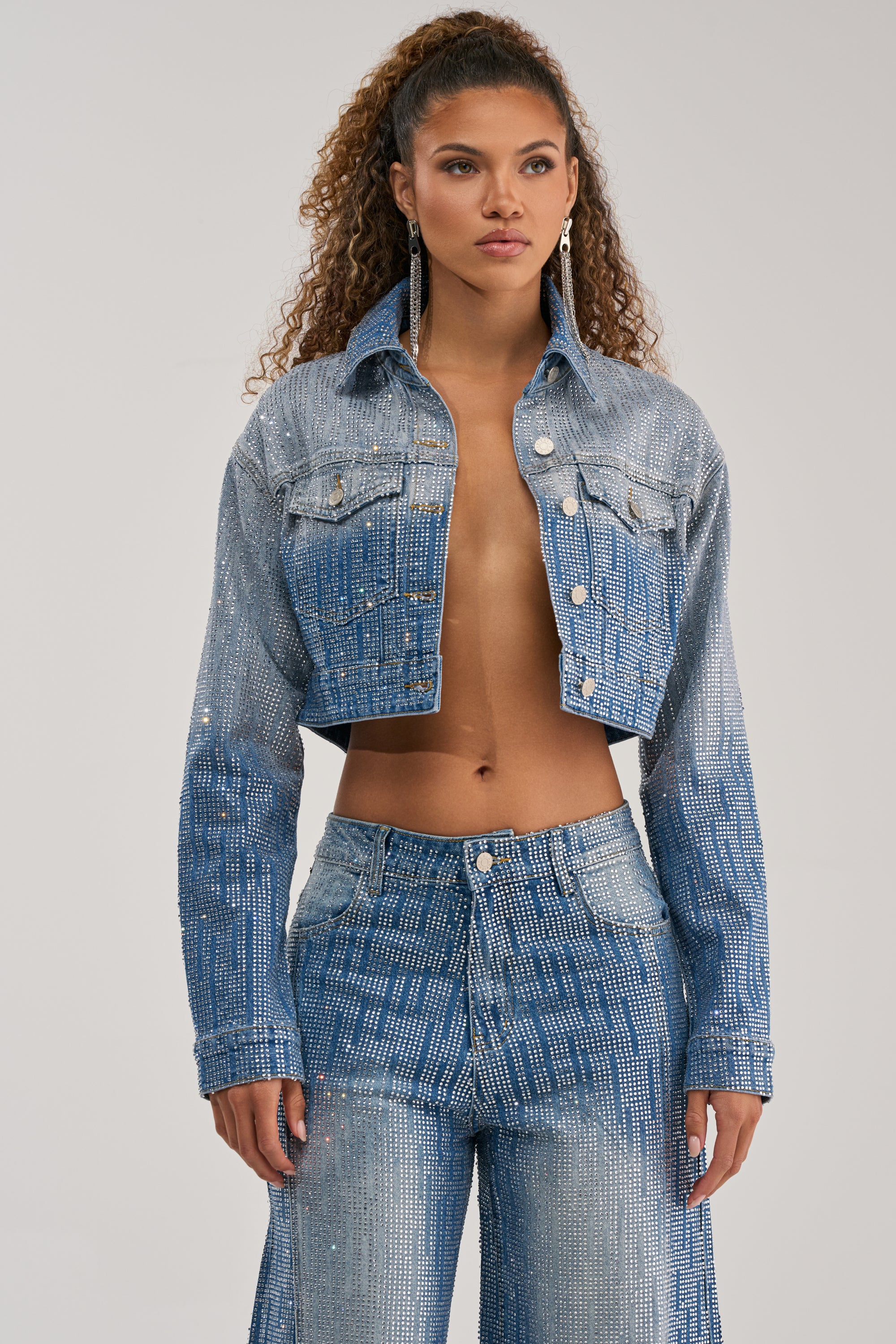 A woman with curly hair and long earrings stands against a plain background, effortlessly showcasing bold streetwear style in the MIND SPINNING EMBELLISHED DENIM JACKET—a cropped ombre denim piece adorned with rhinestone embellishments—paired with matching high-waisted jeans.