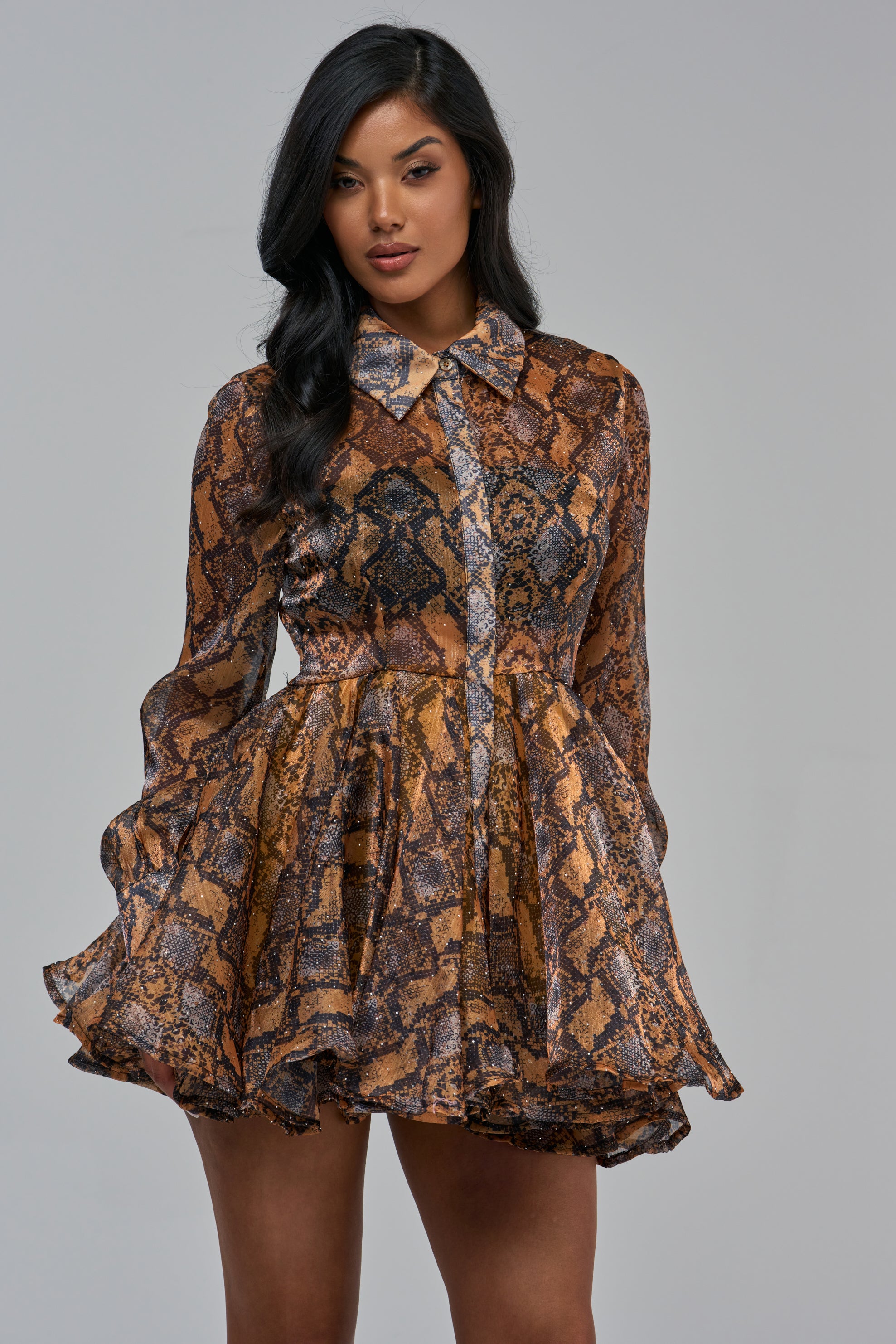 A person wearing a WILD RHYTHM SNAKE PRINT MINI DRESS IN ORANGE MULTI, featuring a long-sleeved, collared, button-down bodice and a flared skirt.