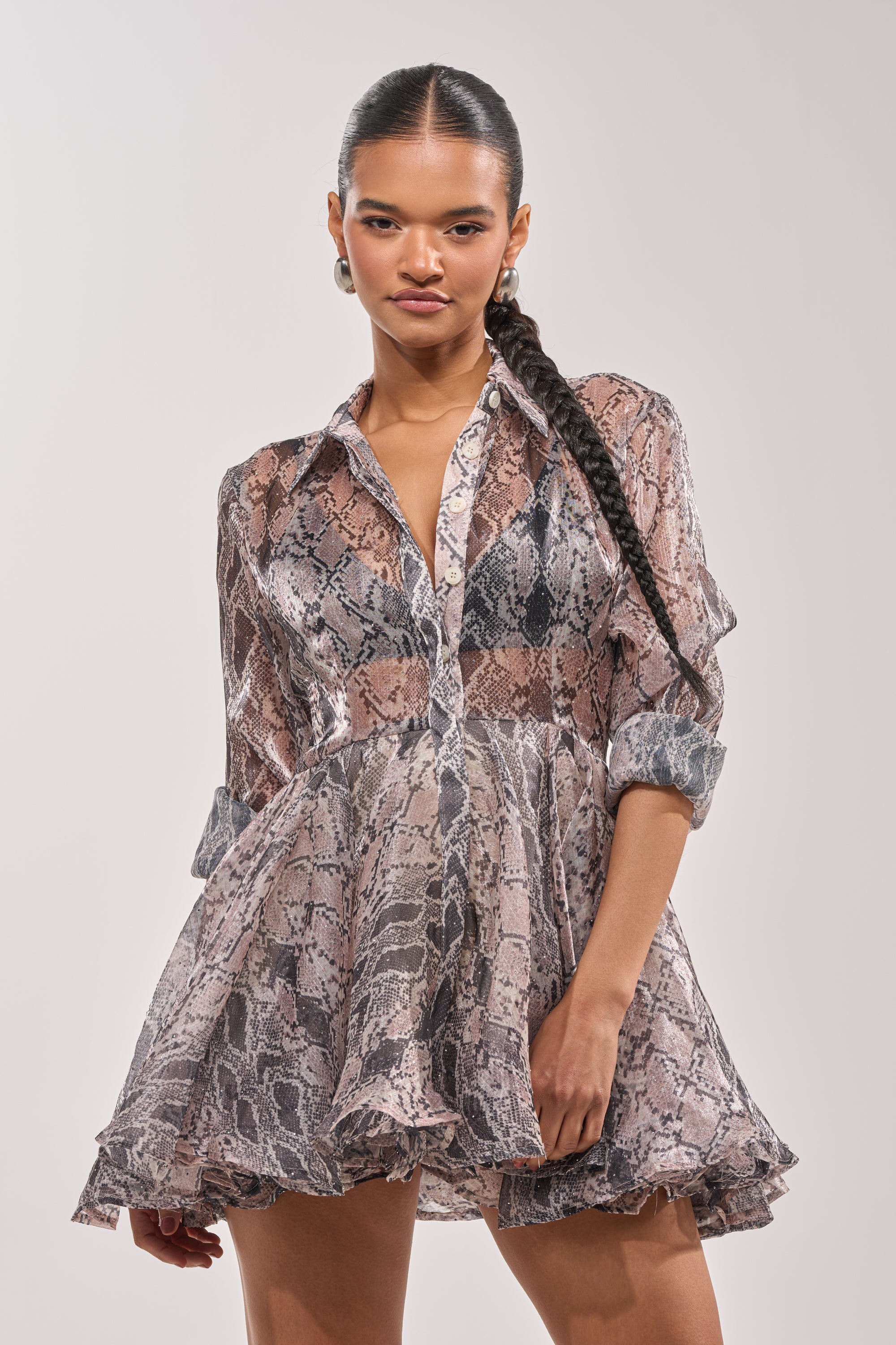 A woman with long, braided hair stands confidently against a plain light background, wearing the WILD RHYTHM SNAKE PRINT MINI DRESS featuring sheer fabric, ruffles, and rolled-up sleeves. 0