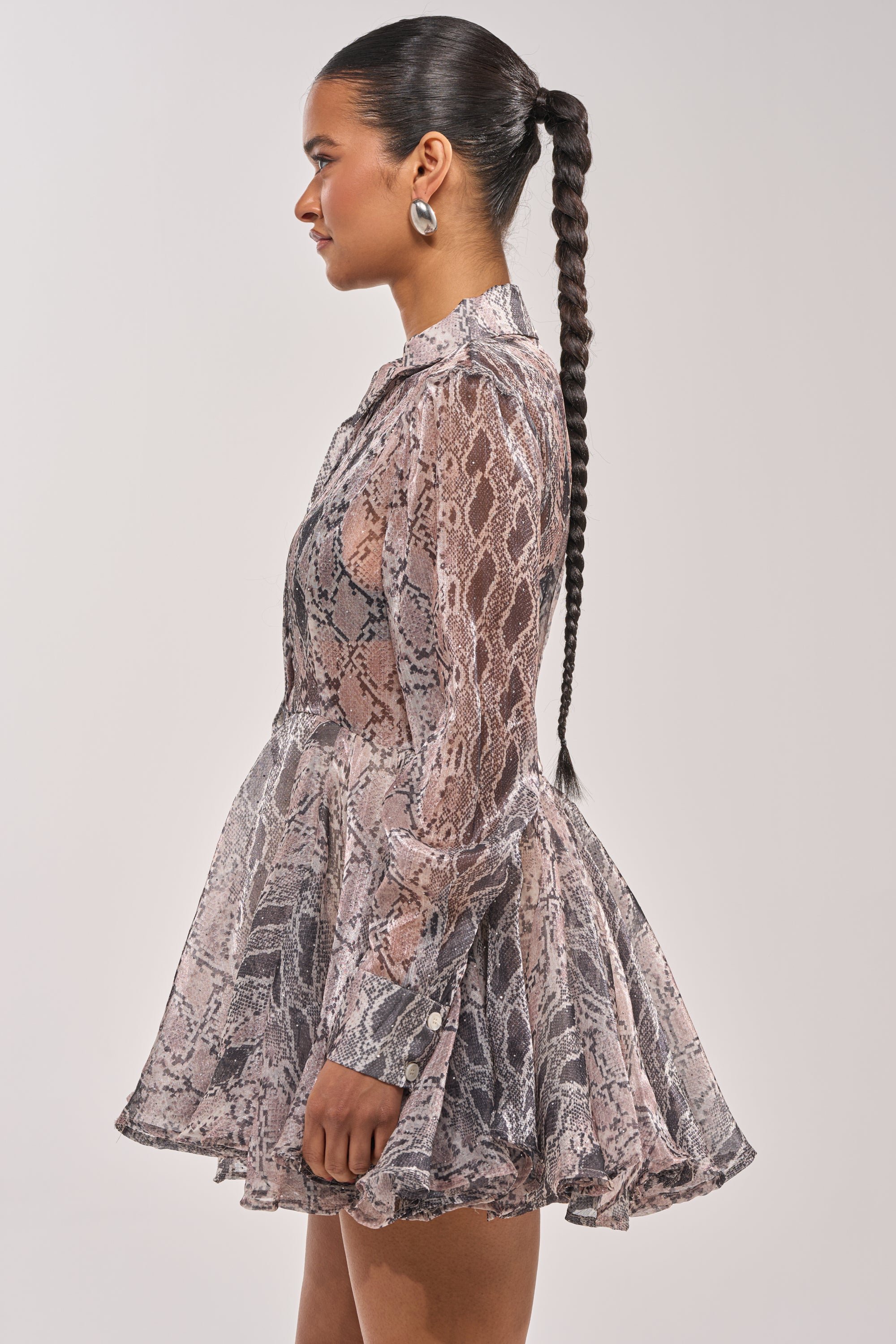 A woman in profile wears the WILD RHYTHM SNAKE PRINT MINI DRESS—a fit-and-flare, long-sleeved pink and gray snakeskin mini. She sports a long braided ponytail and round earrings against a plain background. 3