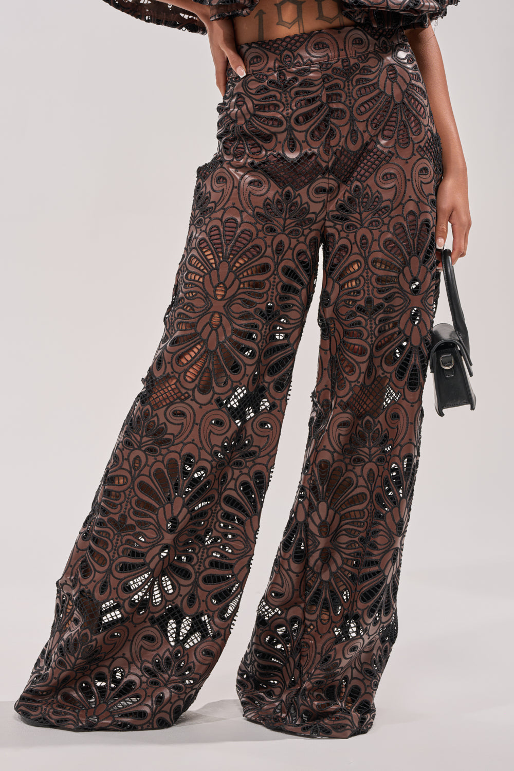 Someone models the LENA LACE AND PU RELAXED PANT, featuring a wide-leg high-rise fit with an intricate brown and black floral lace pattern, while holding a small black handbag against a plain background. 1