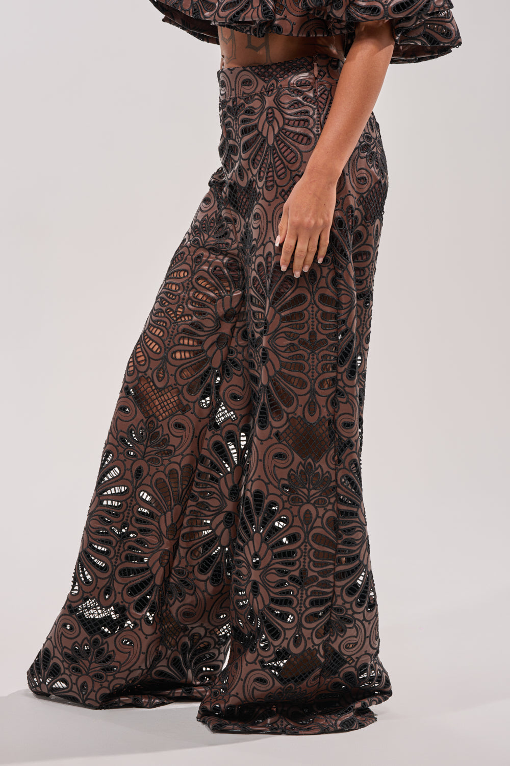 A person models the LENA LACE AND PU RELAXED PANT—wide-leg, high-waisted brown faux leather pants with black lace accents—against a plain light gray background, showcasing the design's elegant and stylish appeal. 3