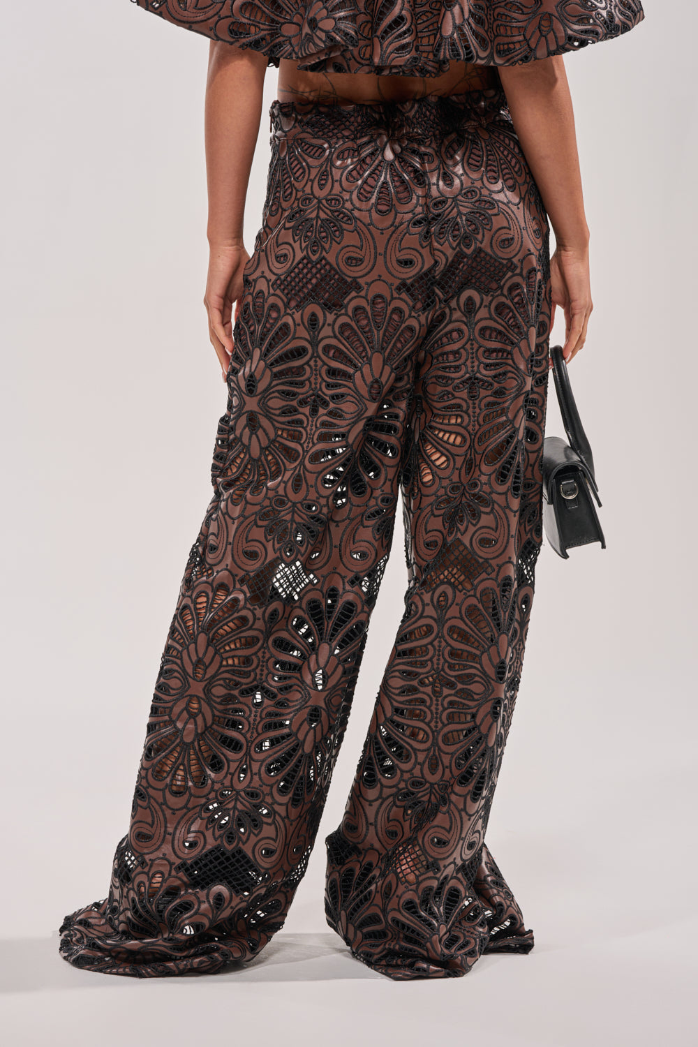 A person is seen from the back wearing LENA LACE AND PU RELAXED PANT—high-waisted, wide-leg pants with a black and brown floral pattern and sequin details—paired with a small black handbag against a plain background. 4