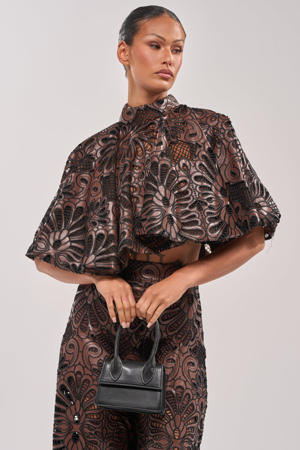 A woman models the LENA LACE AND PU BLOUSE, a sheer black and brown evening top with floral patterns and lace details, holding a small black handbag against a plain light gray background. 0