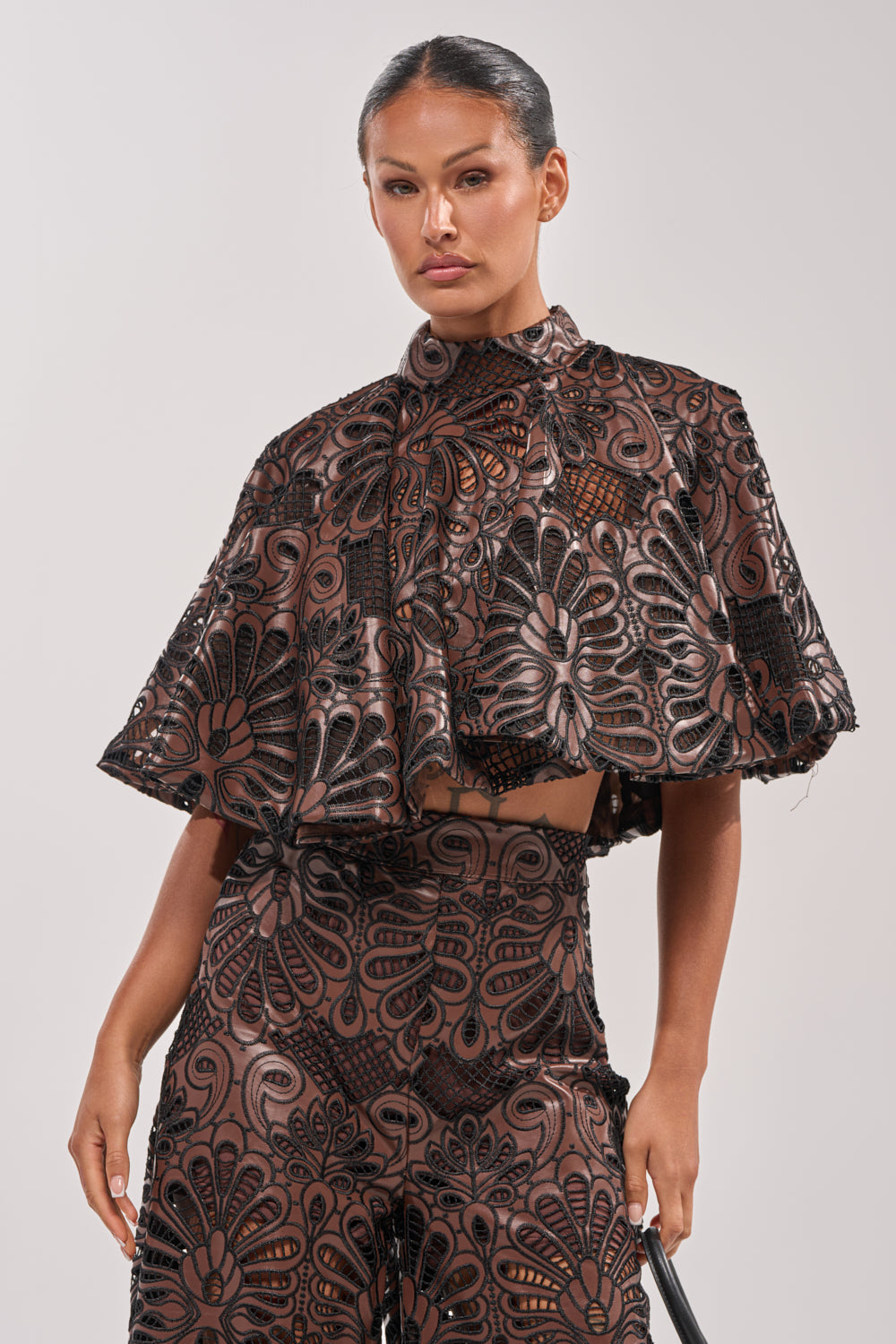 A woman poses in the LENA LACE AND PU BLOUSE, a high-neck, short-sleeve crop top with lace details and black and brown floral cutouts, paired with matching high-waisted pants. She wears her hair slicked back against a plain light background. 1