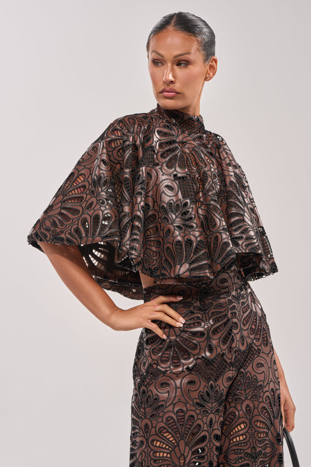 A woman stands against a plain light background, confidently wearing the LENA LACE AND PU BLOUSE—a cropped faux leather top with shiny fan-like designs—paired with matching high-waisted pants for a striking evening look. 2