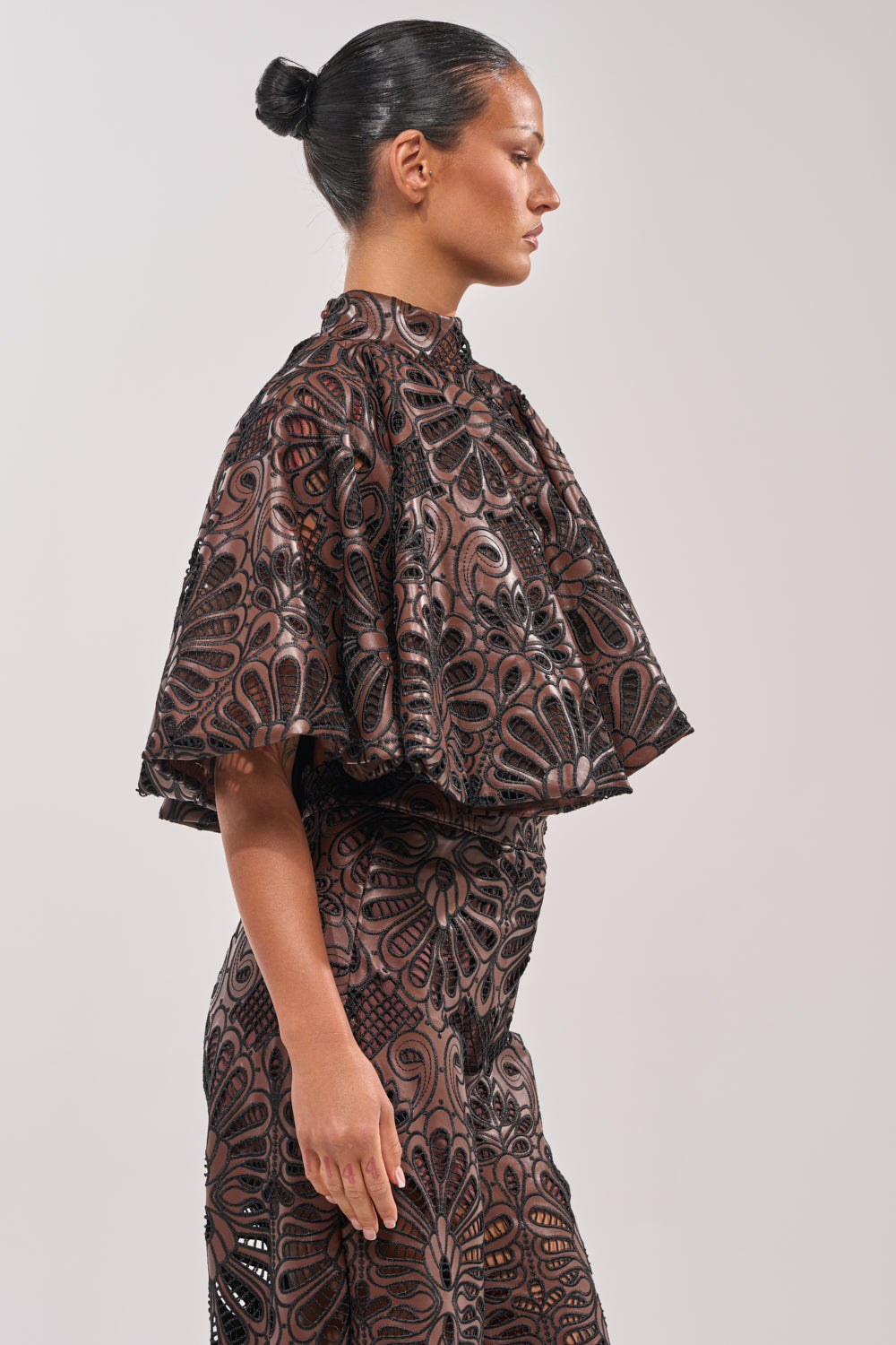 A woman with dark hair in a sleek bun models the LENA LACE AND PU BLOUSE—a cropped faux leather top with lace and floral details, featuring wide cape-like sleeves, styled elegantly for evening events. She poses in profile against a plain background. 3