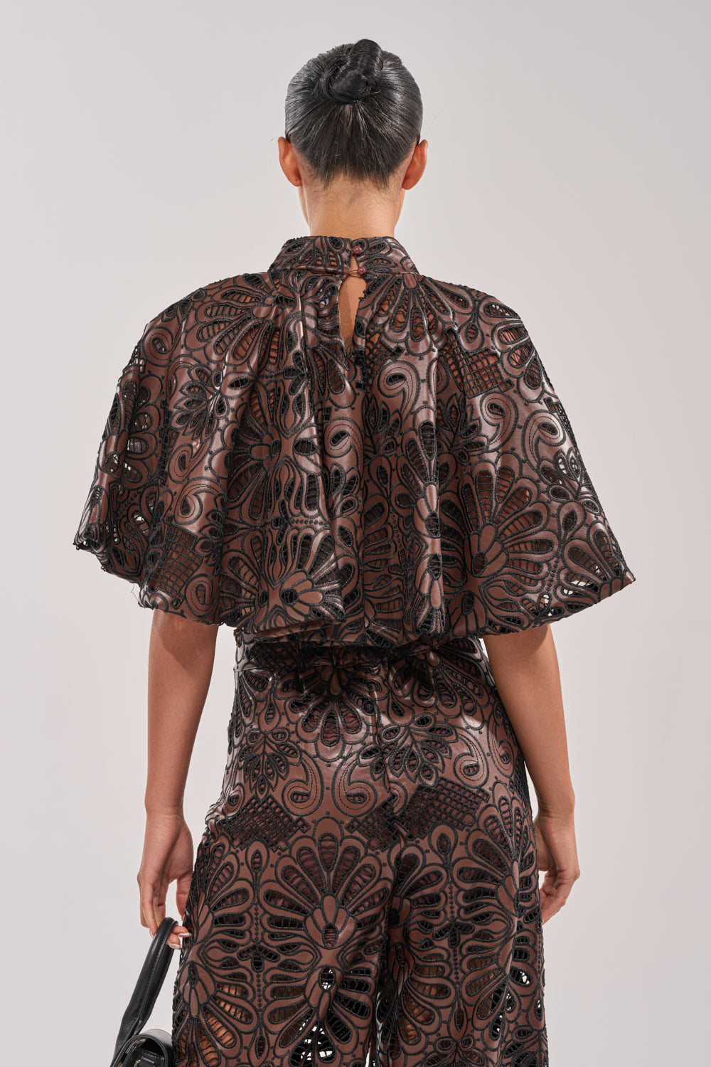 Seen from the back, a person with dark hair in a sleek bun wears the LENA LACE AND PU BLOUSE featuring intricate floral patterns and lace details—perfect for evening events—paired with patterned pants and holding a small black bag. 4