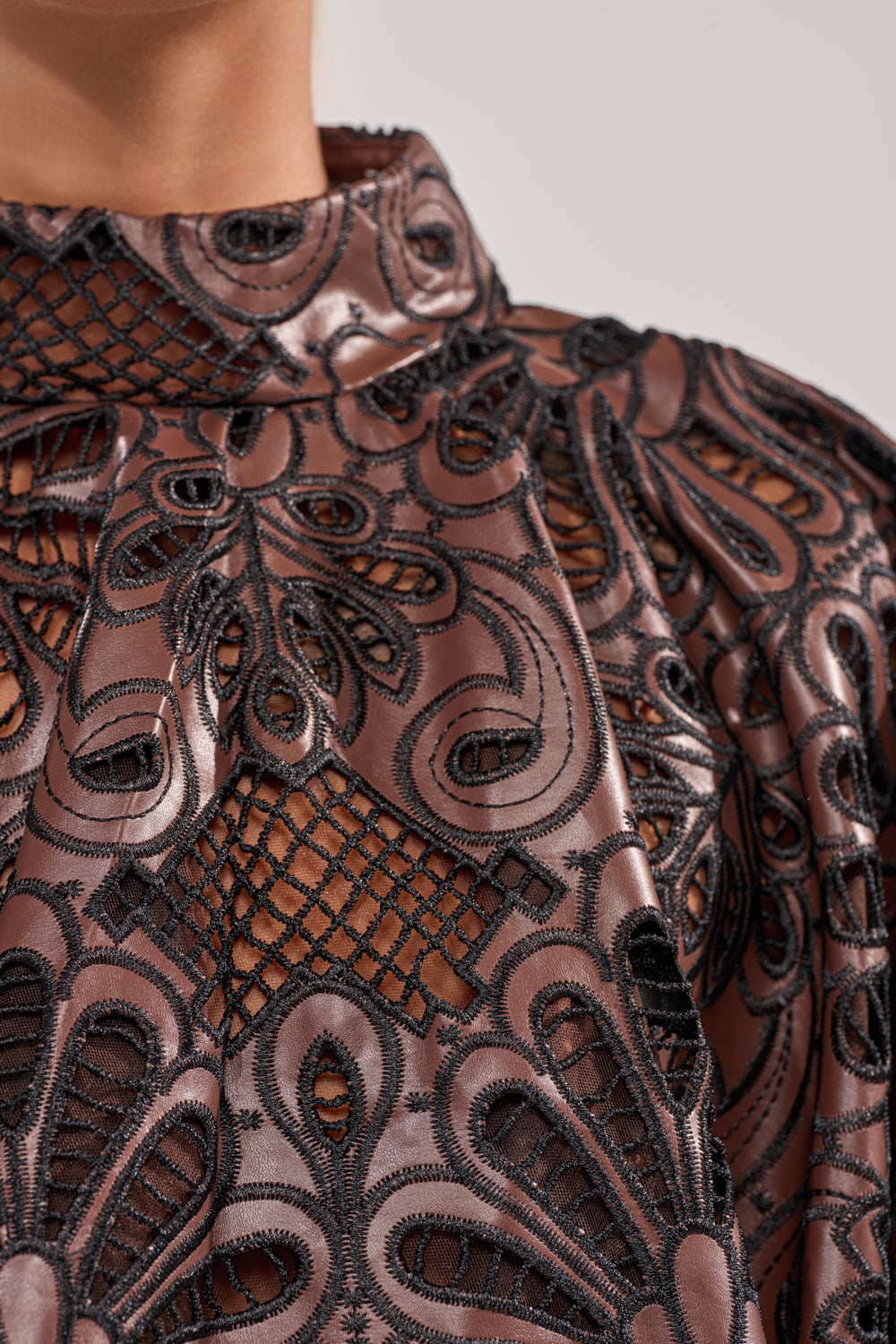 Close-up of a person wearing the LENA LACE AND PU BLOUSE, featuring a high neck, metallic brown shine, black lace with floral and abstract patterns, sheer panels, and a unique textured layered look. 7