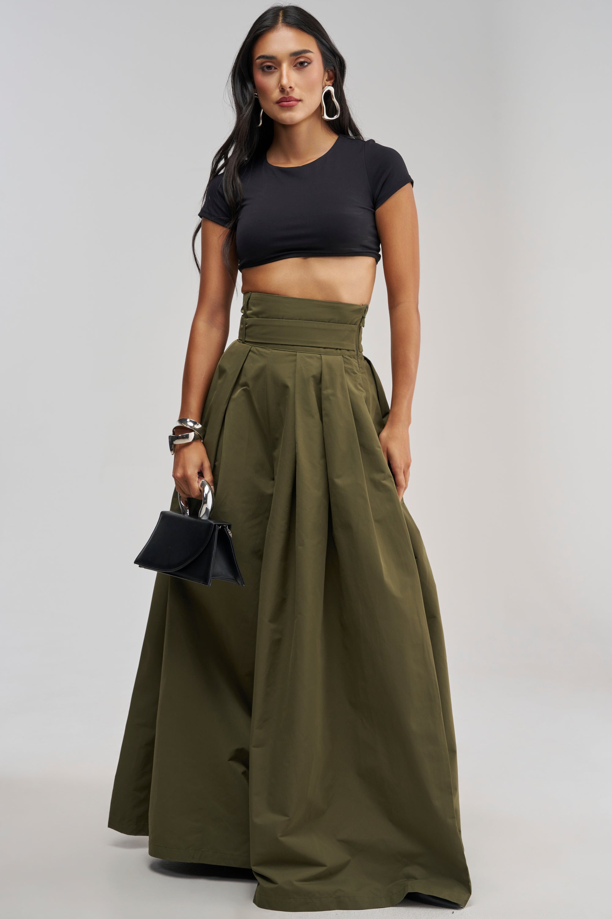 A woman wearing a black crop top and the DISCO DOLL TROUSER IN OLIVE, styled with hoop earrings, bracelets, and a small black purse, stands against a plain light background.
