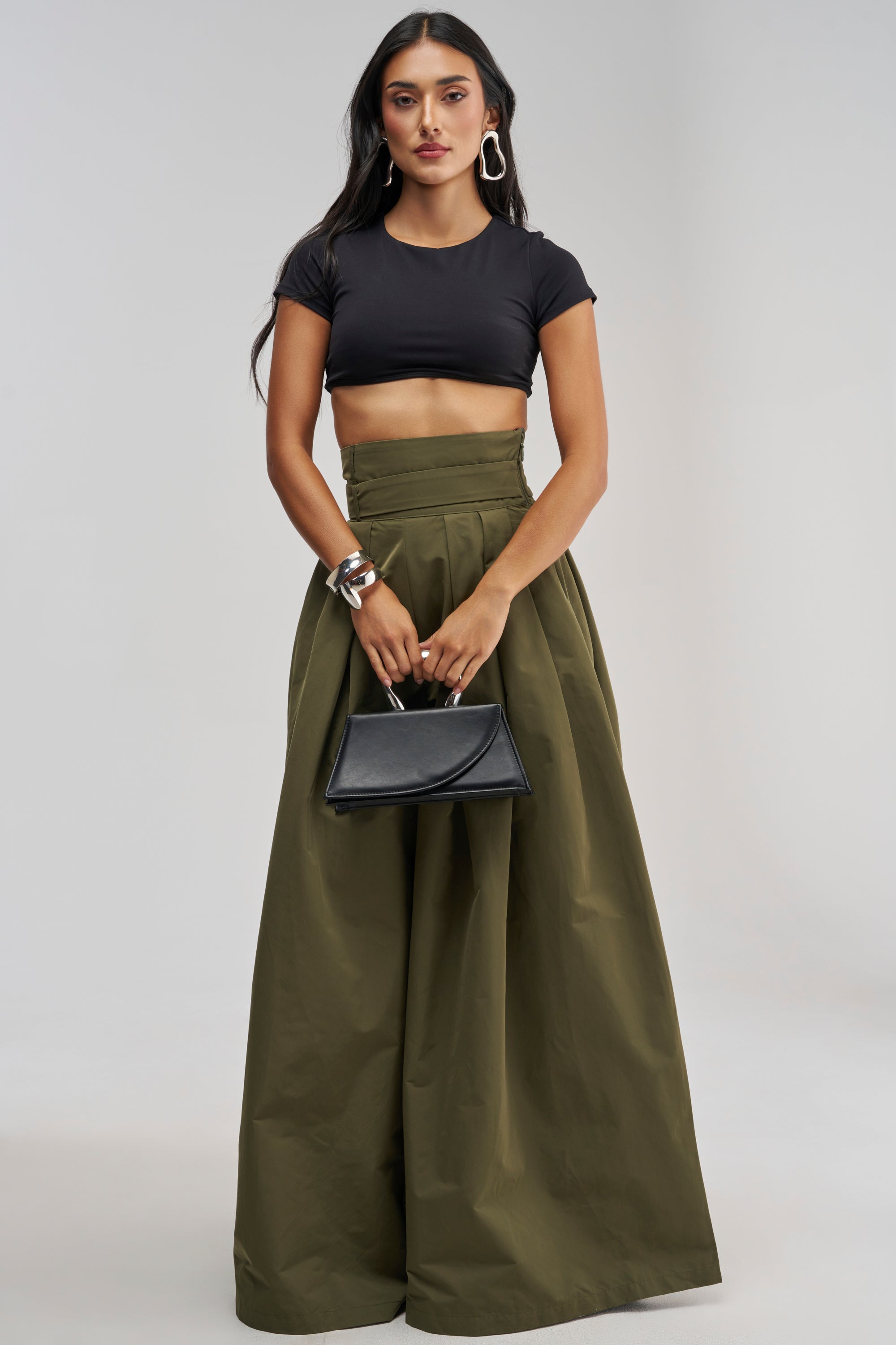 A woman wearing the DISCO DOLL TROUSER IN OLIVE, styled with a black crop top and a small black handbag, showcases long dark hair, hoop earrings, and silver bangles. Posed against a plain light background, she effortlessly blends casual chic with polished office-wear style.