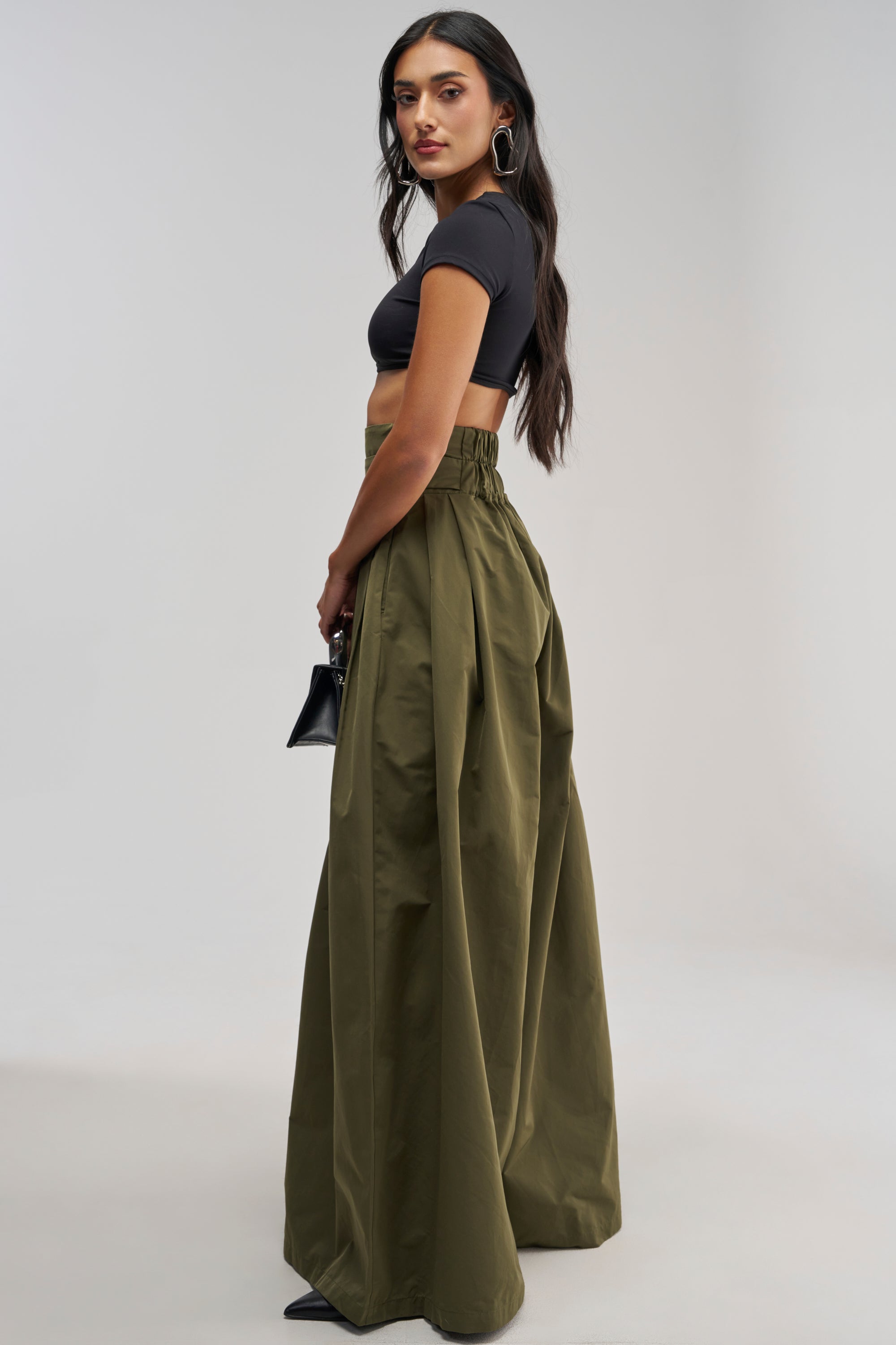 Styled for the office, this woman is wearing a black crop top paired with the DISCO DOLL TROUSER IN OLIVE, black heels, hoop earrings, and carrying a small black handbag as she stands against a plain light background.