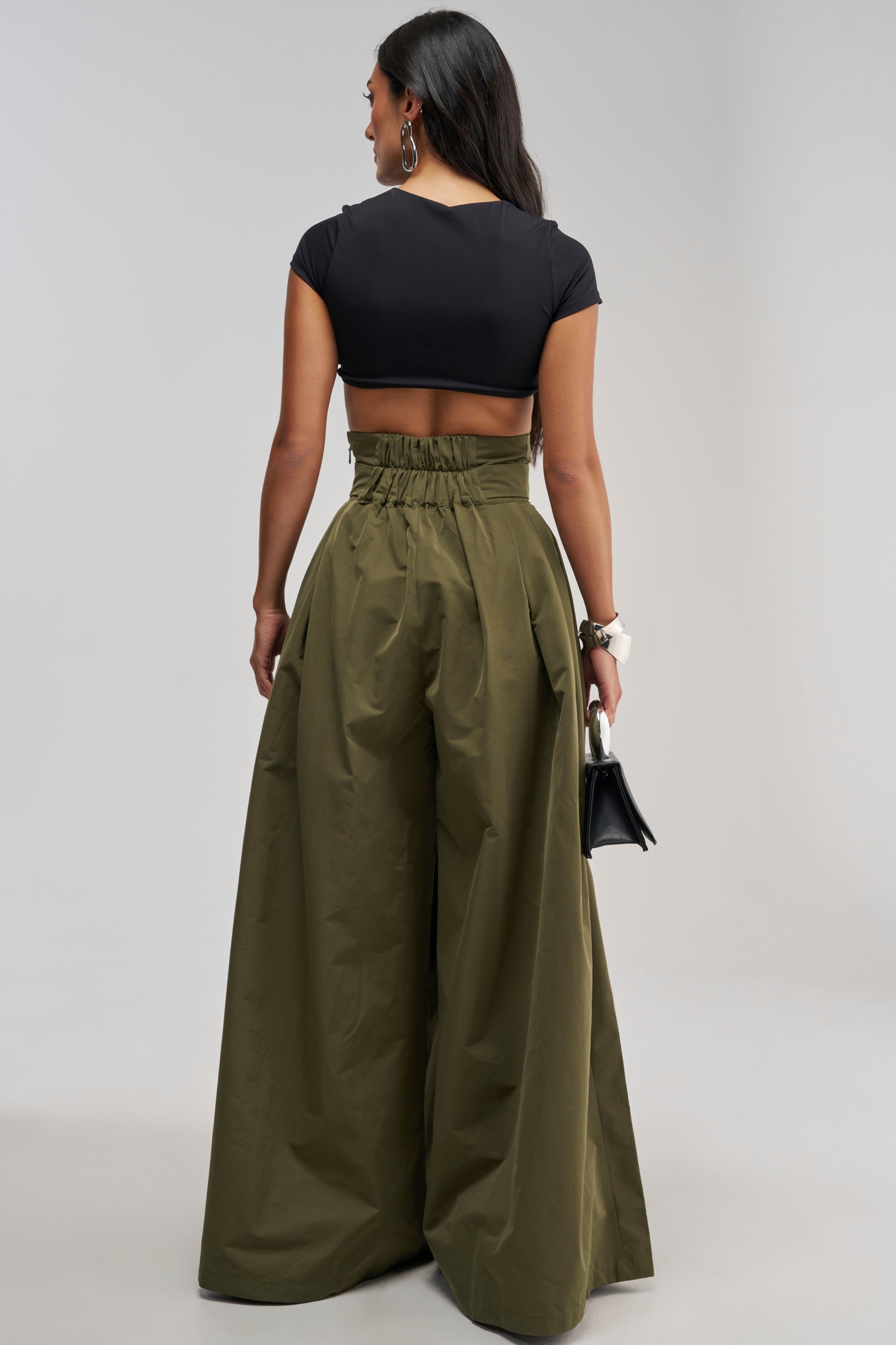 A woman stands facing away, wearing a black crop top and the DISCO DOLL TROUSER IN OLIVE—high-waisted, wide-leg trousers. She holds a small black handbag and has long dark hair, creating a chic office-wear look.