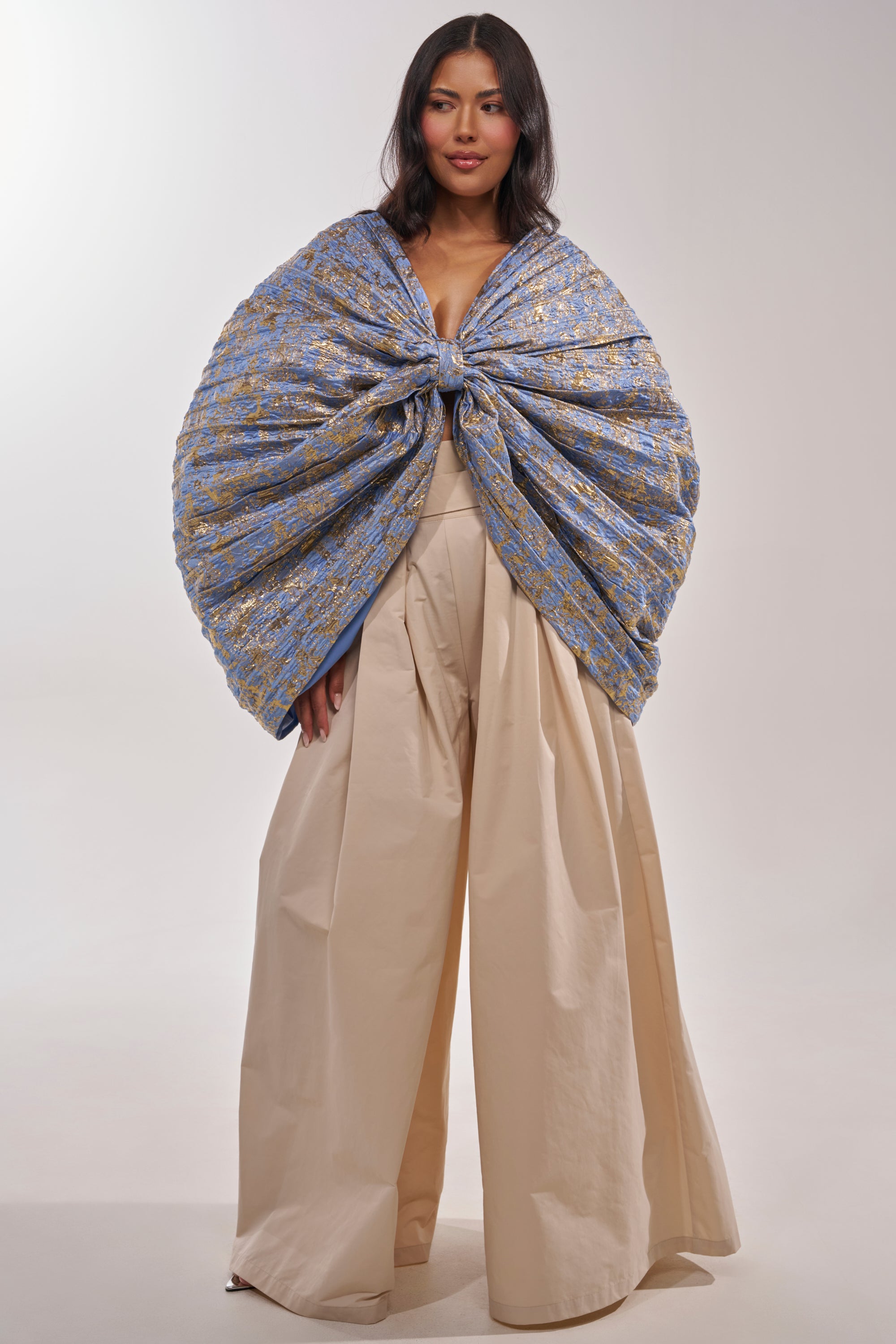 A woman with long dark hair and a confident expression stands against a plain background wearing DISCO DOLL TROUSER in beige, paired with a dramatic blue cape featuring gold details knotted at her chest. 7