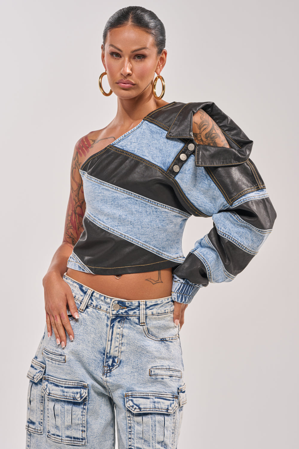 A tattooed woman wears large hoops and the DISILLUSIONED DENIM PU TOP—a unique off-shoulder denim top with black leather patches—paired with light wash cargo jeans, standing against a plain background. 0