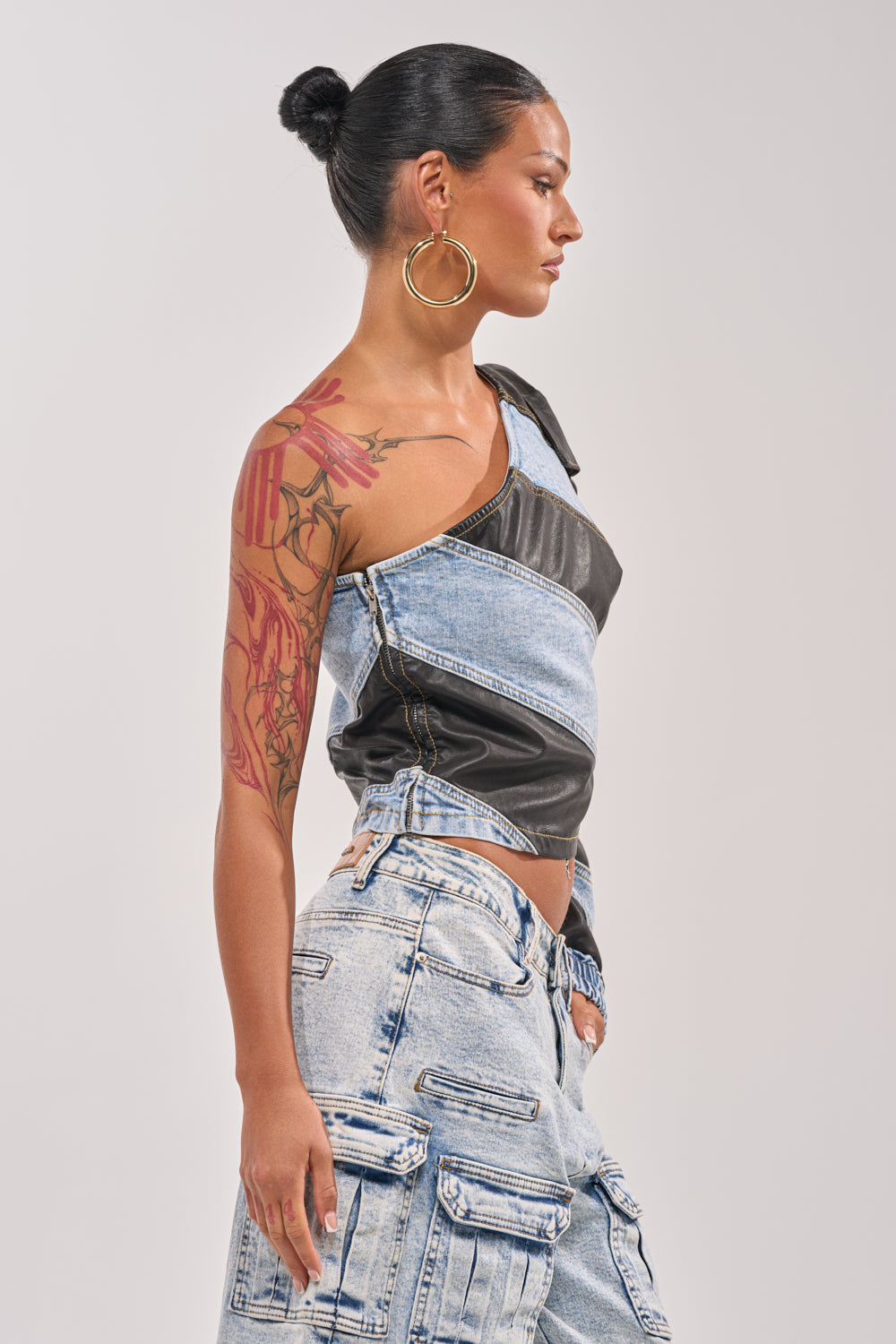 A woman with her dark hair in a bun, large hoop earrings, and a colorful floral tattoo on her upper arm stands in profile wearing the DISILLUSIONED DENIM PU TOP—a one-shoulder mixed denim and leather piece—paired with light-wash cargo jeans. 2