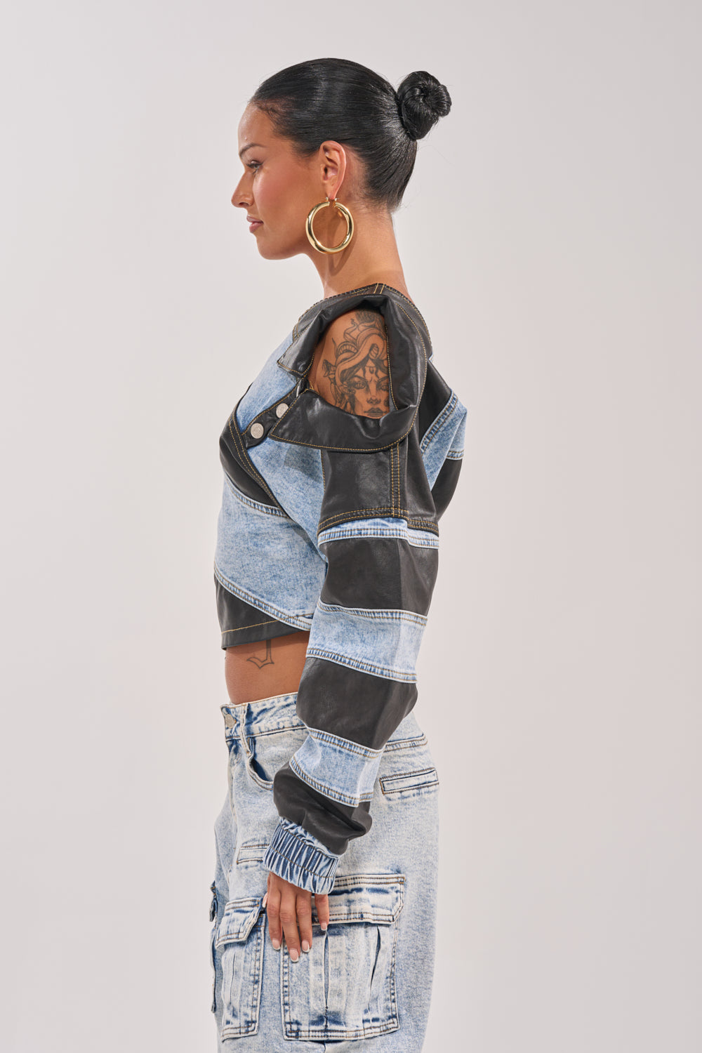 Model with a tattooed arm wears the DISILLUSIONED DENIM PU TOP, styled with large hoop earrings and light-wash denim cargo jeans, standing in profile against a plain light background. 3
