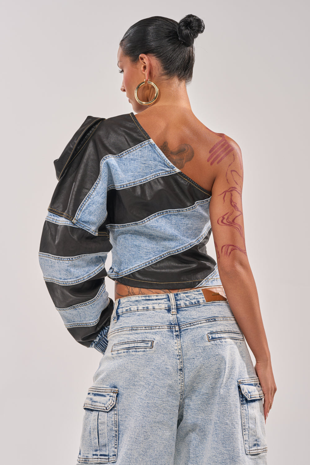 A woman with her hair in a bun and large hoop earrings wears the DISILLUSIONED DENIM PU TOP, a mixed denim asymmetrical style with black fabric strips, paired with light denim cargo pants. Red abstract tattoos decorate her shoulder and arm. 4