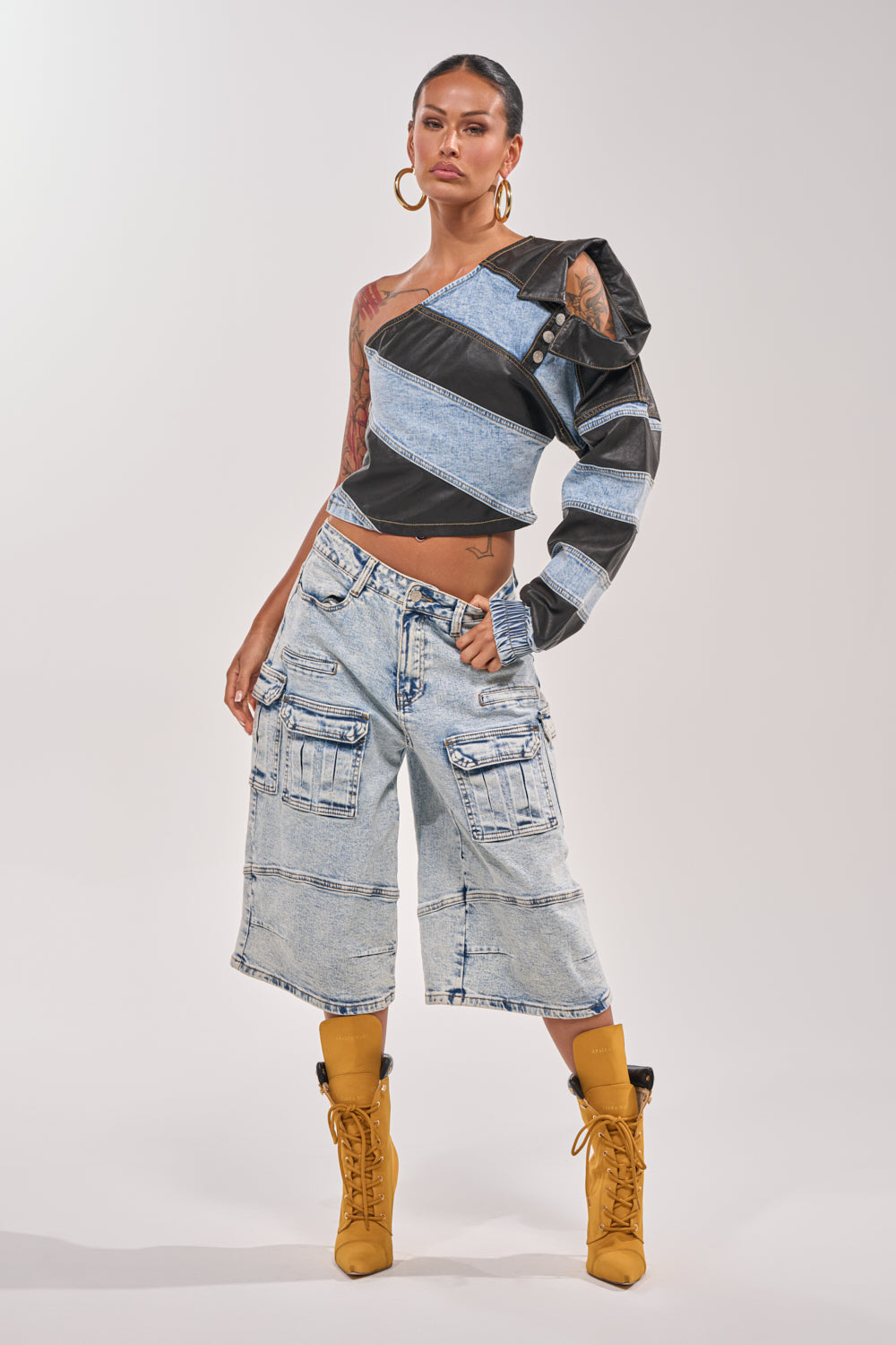 A person stands confidently in the DISILLUSIONED DENIM PU TOP with one sleeve, paired with light-wash denim cargo shorts, mustard yellow lace-up boots, hoop earrings, and showcasing visible arm tattoos. 5