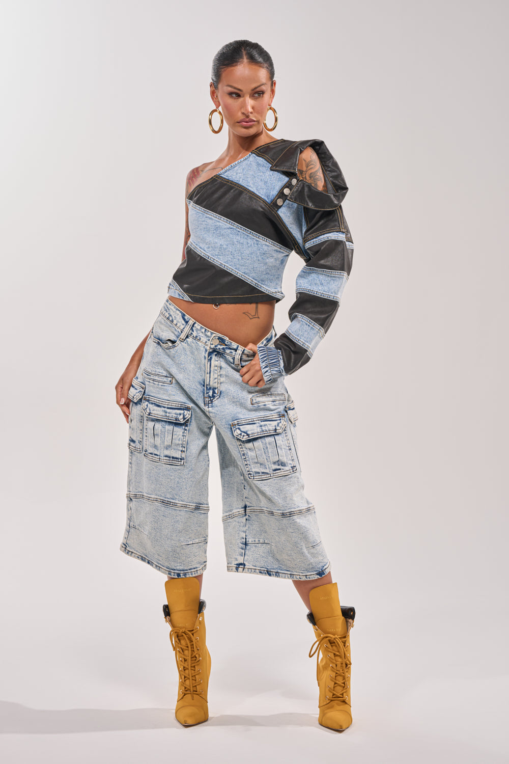 A woman poses confidently against a white background wearing the DISILLUSIONED DENIM PU TOP with a colorblock design, paired with light denim cargo knee-length shorts, tan lace-up boots, and large hoop earrings. 6