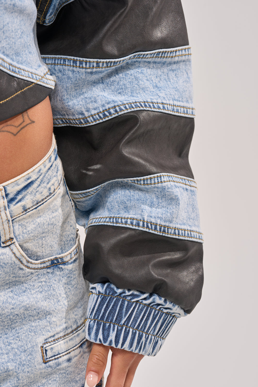 A close-up of a person wearing the DISILLUSIONED DENIM PU TOP, featuring mixed denim and black leather-like striped sleeves; a tattoo is visible at the waist and their hand rests by their side. 8