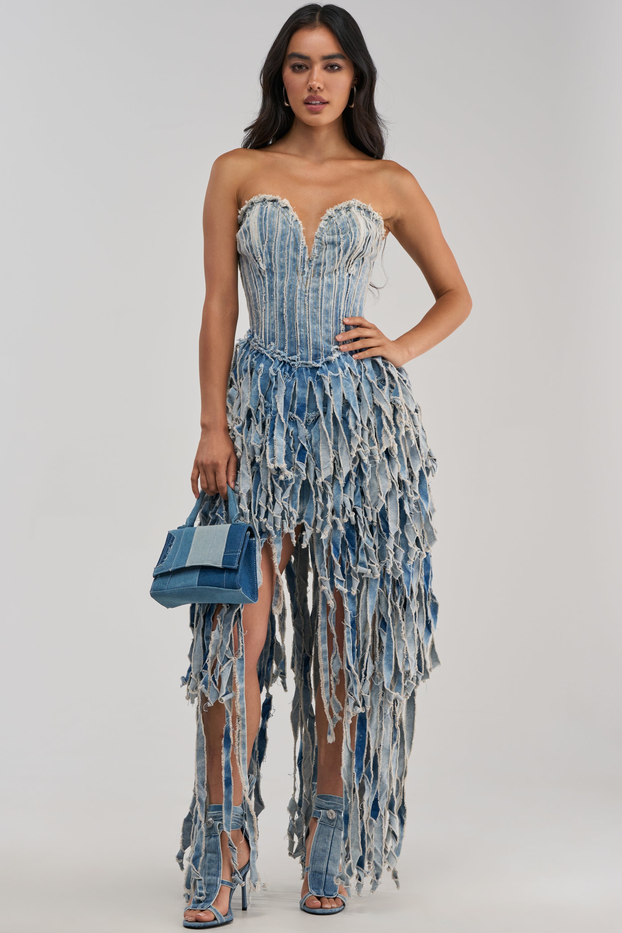 A woman wears the HERE FOR A GOOD TIME STRAPLESS DENIM DRESS, featuring distressed details and a frayed, jagged hem reminiscent of a fringe high-low silhouette. She pairs the dress with matching denim heels and a small denim handbag, standing against a plain light background.