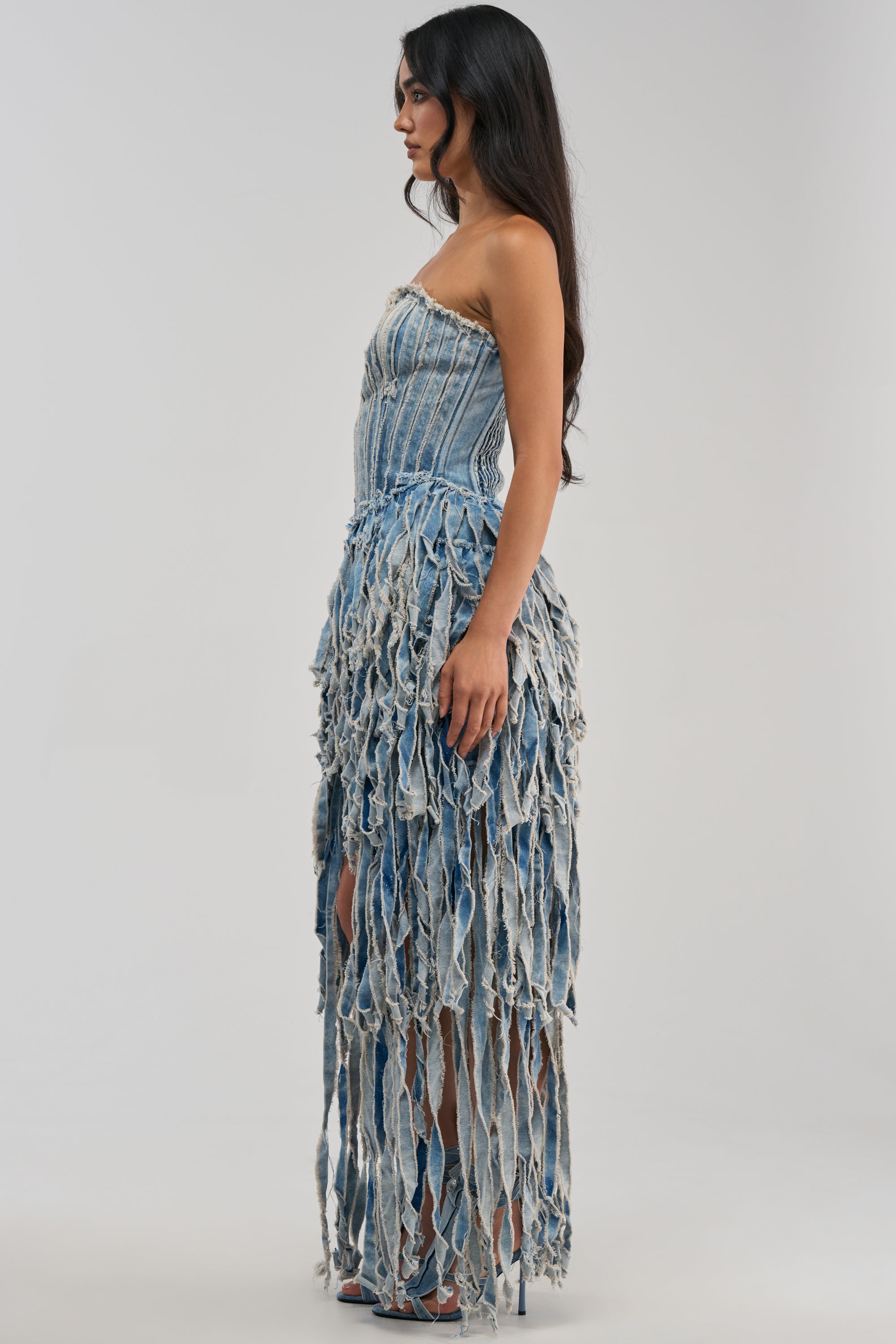 A woman with long dark hair stands in profile against a plain background, wearing the HERE FOR A GOOD TIME STRAPLESS DENIM DRESS, which features a fitted bodice and a skirt made of long, frayed denim strips.